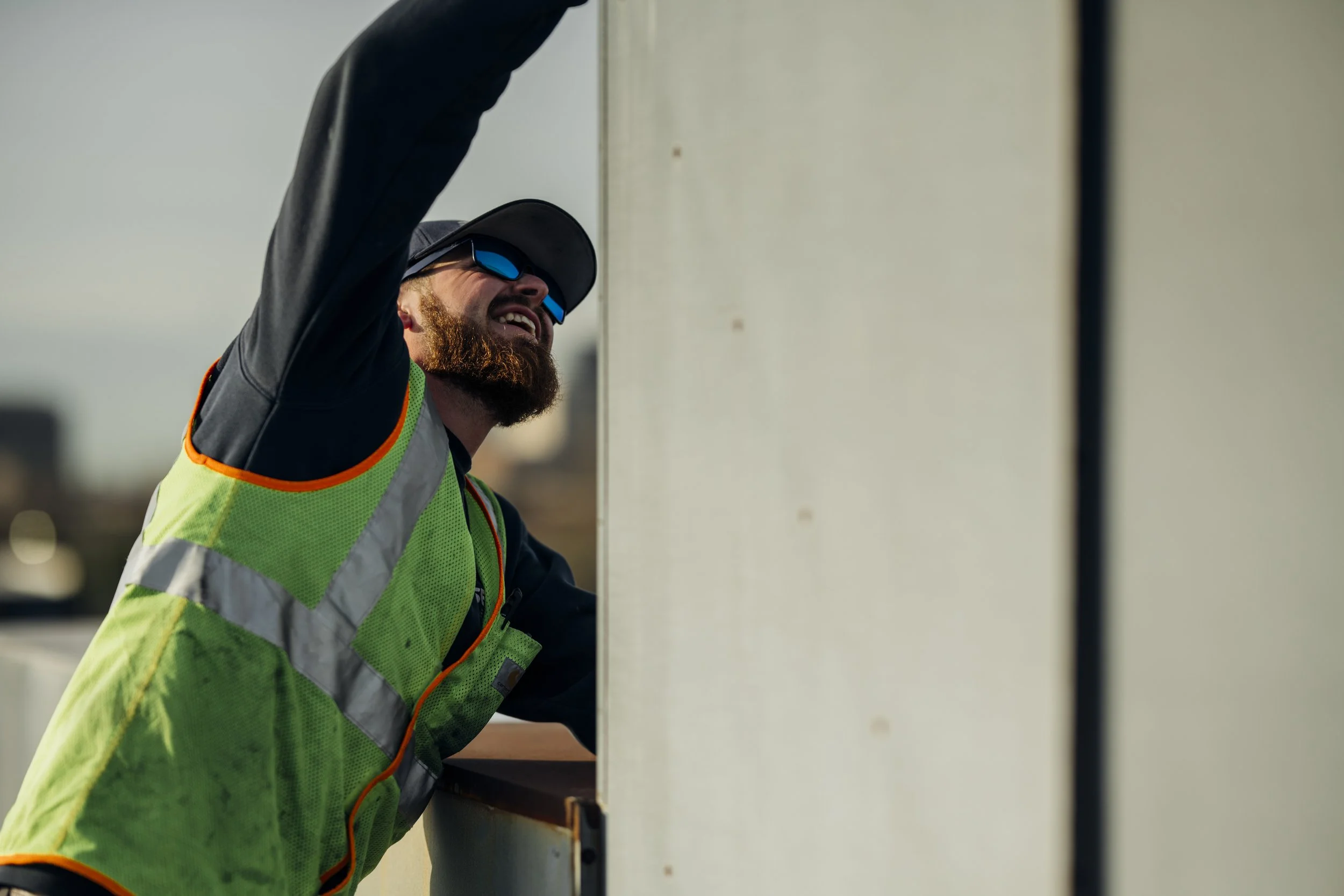 A worker with a beard, wearing sunglasses, a hard hat, and a high visibility safety vest, is smiling and leaning on a large white structure outdoors.