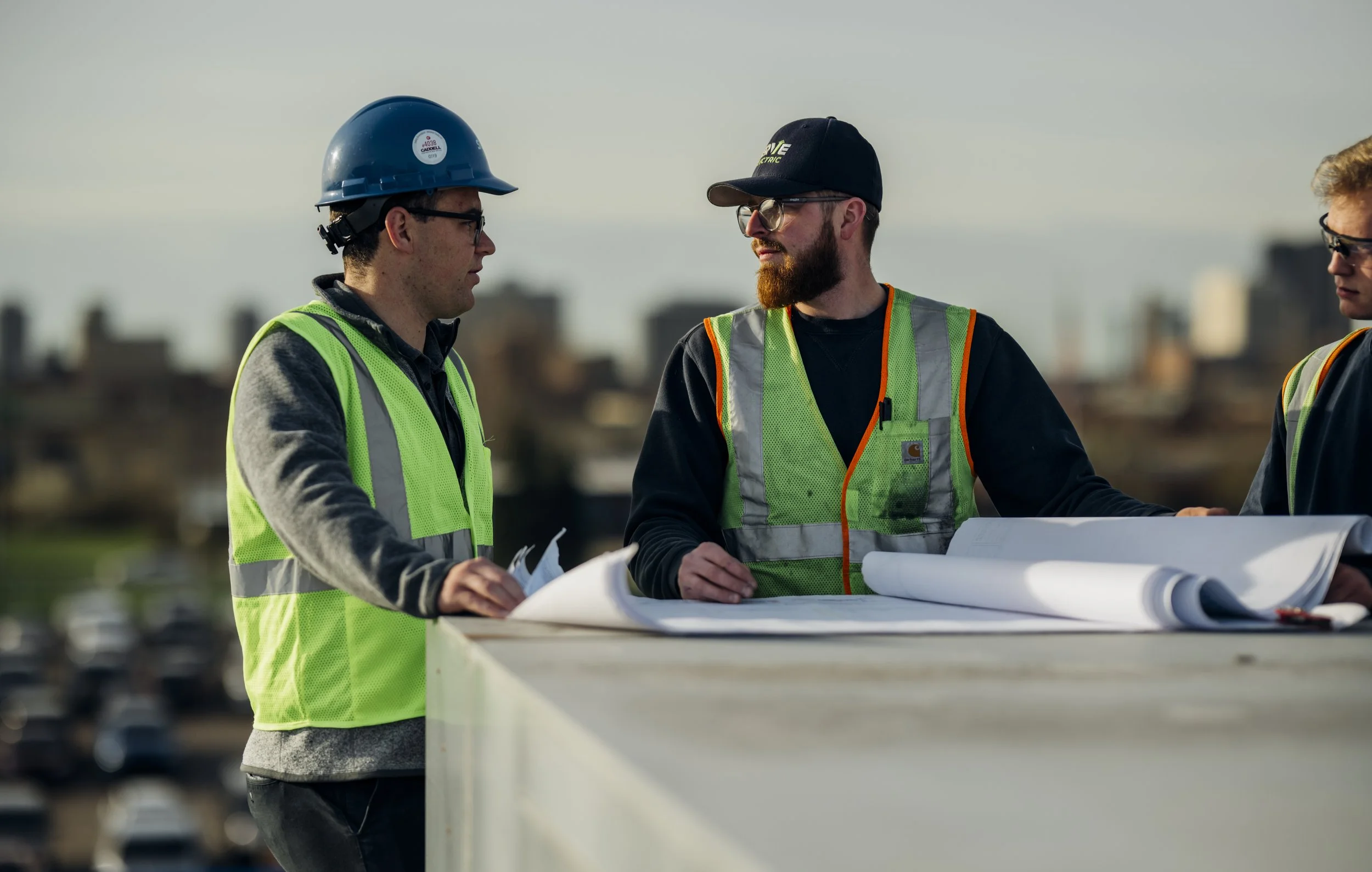 Three construction workers wearing safety vests and hard hats are discussing plans outdoors with a city skyline in the background.