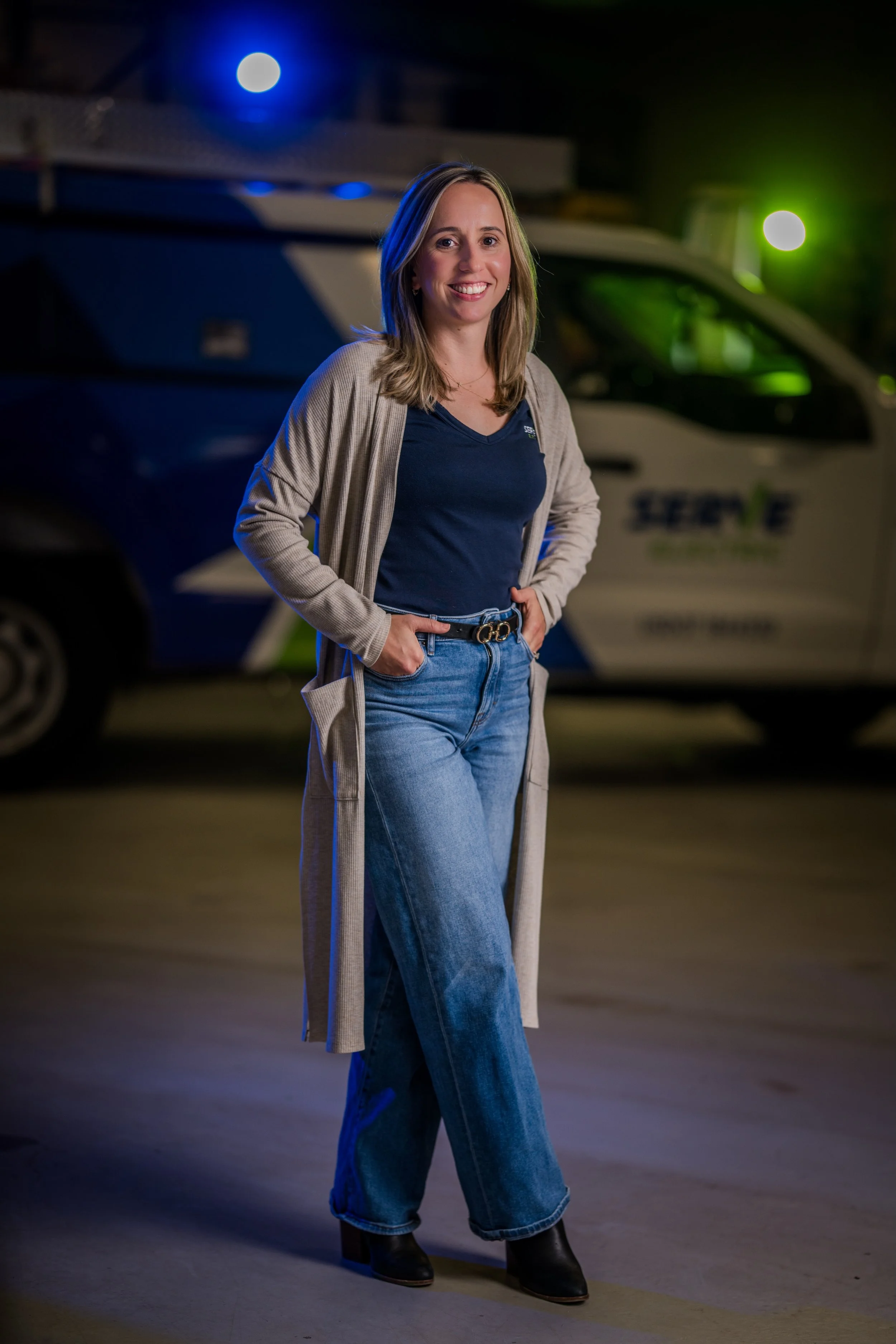A woman stands outdoors at night in front of a police truck, smiling and wearing a casual outfit with a long beige cardigan, navy t-shirt, blue jeans, and black boots.