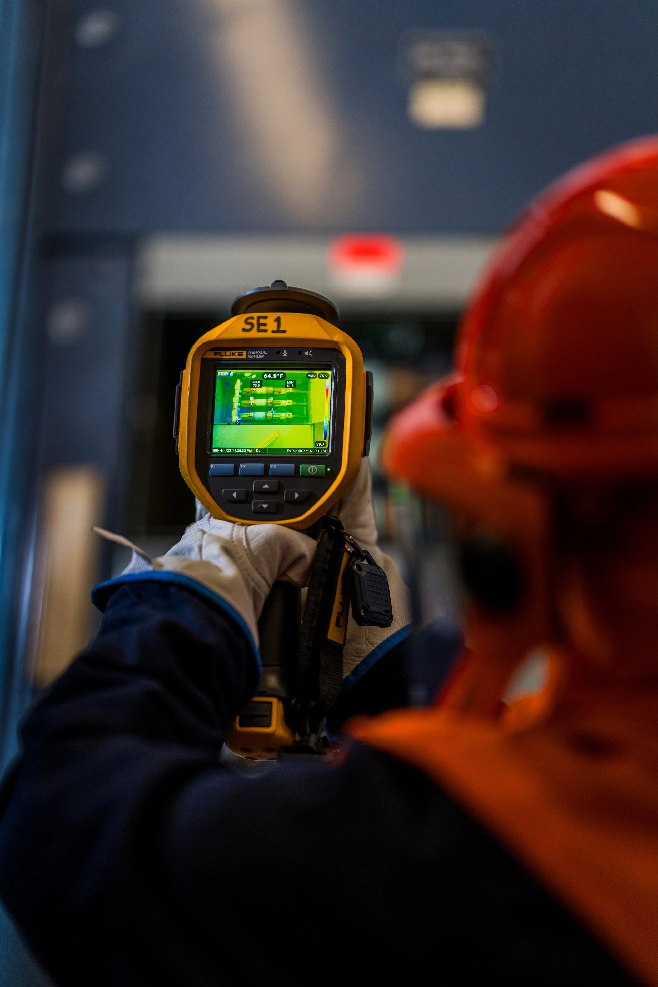 Person wearing gloves holding a thermal imaging device that displays a thermal map, with a partially visible person in an orange jacket in the foreground and a blurry background.