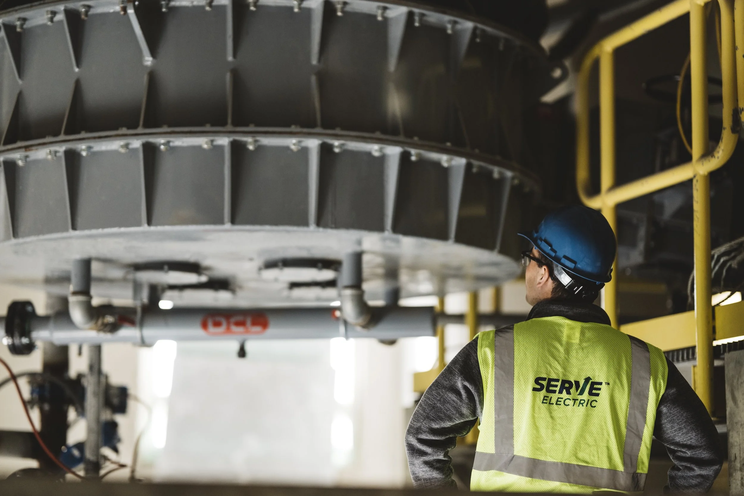 A worker wearing a blue safety helmet, glasses, and a yellow high-visibility vest standing inside an industrial facility, looking at large machinery overhead.