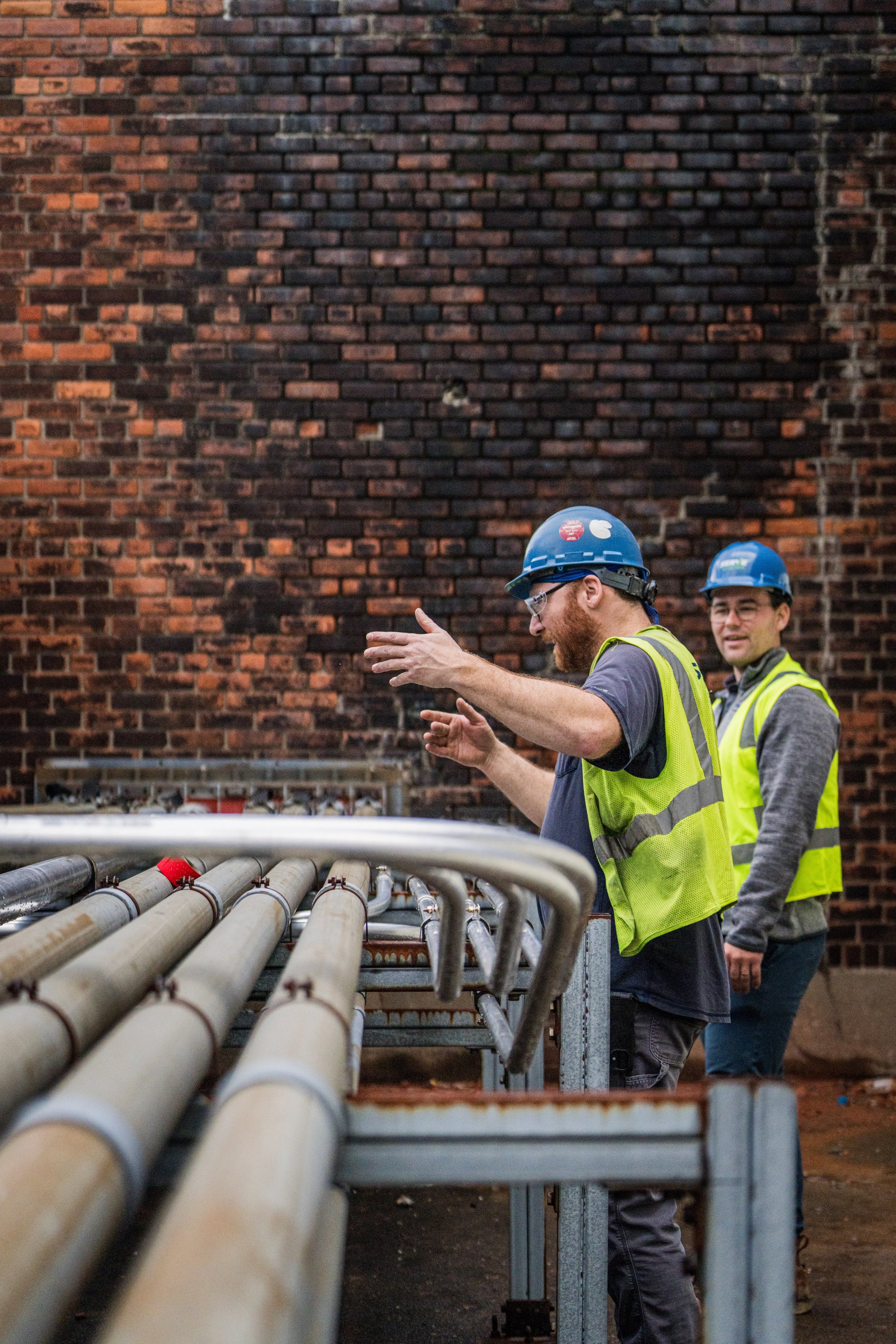 Two construction workers wearing safety helmets and high-visibility vests standing next to a series of metal pipes, with a red brick wall in the background.