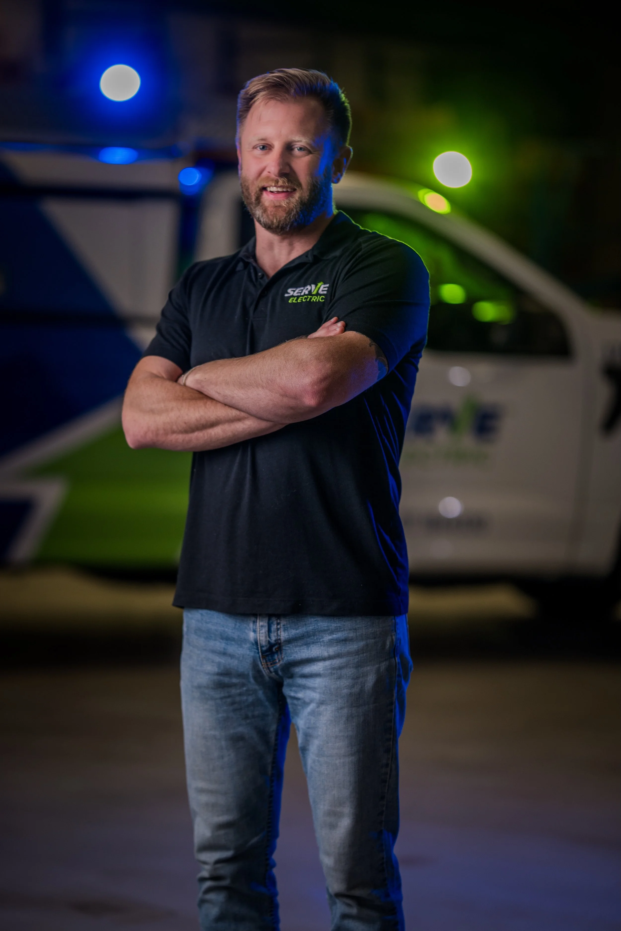 A man with a beard and short hair standing with arms crossed in front of a service vehicle, wearing a black polo shirt with a logo that says 'SERVE ELECTRIC'.
