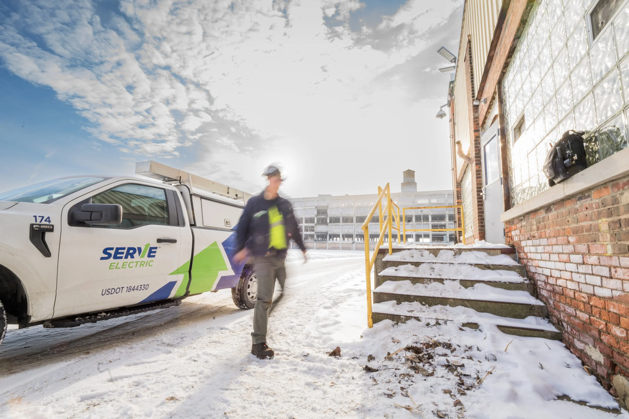 A worker in a yellow safety vest and helmet walking on snow-covered ground near a SERVE Electric vehicle parked outside a brick building with snow on the stairs and ground.