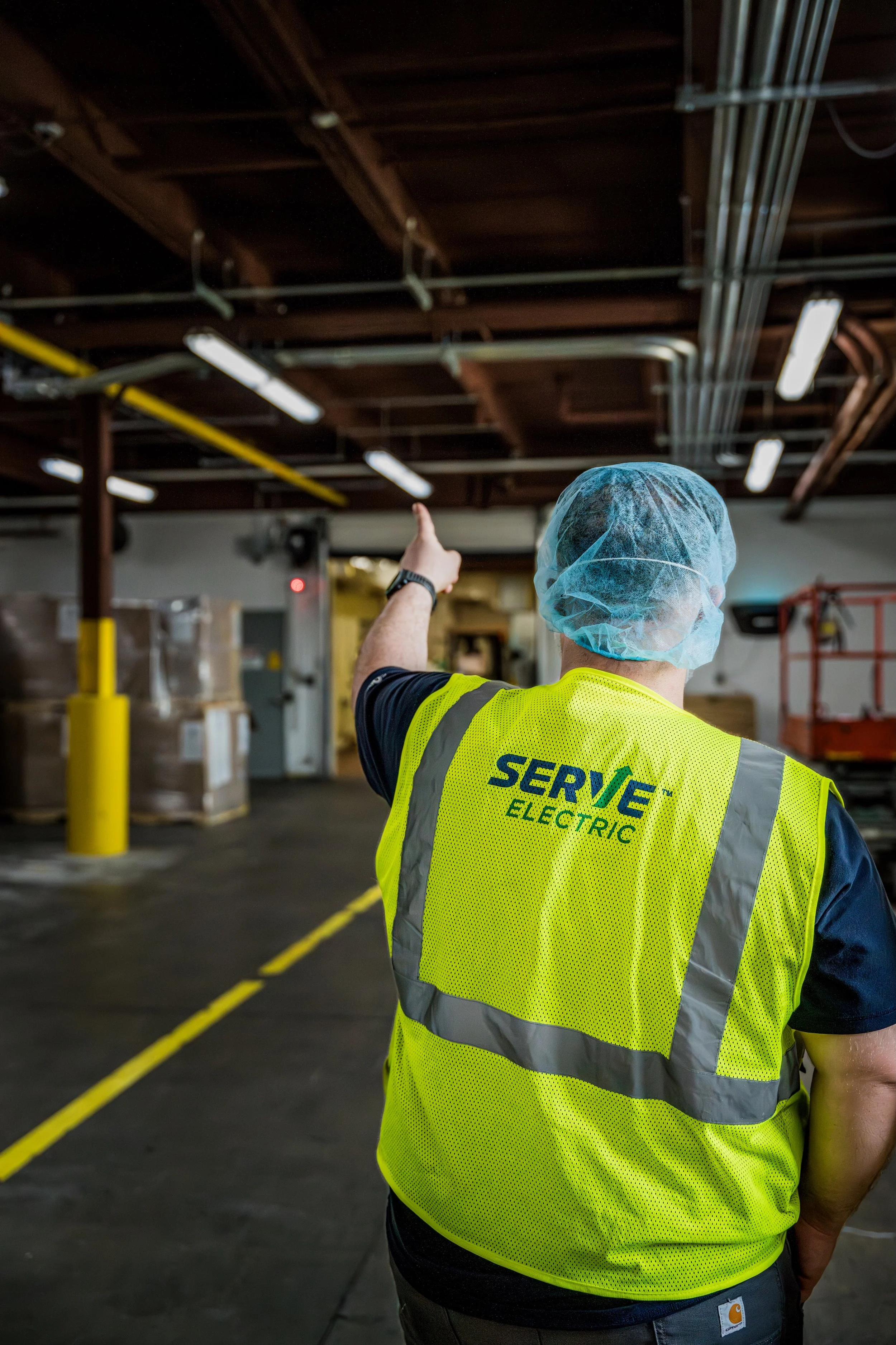 A worker in a yellow safety vest and blue hair net points while inspecting an industrial warehouse or electrical facility.