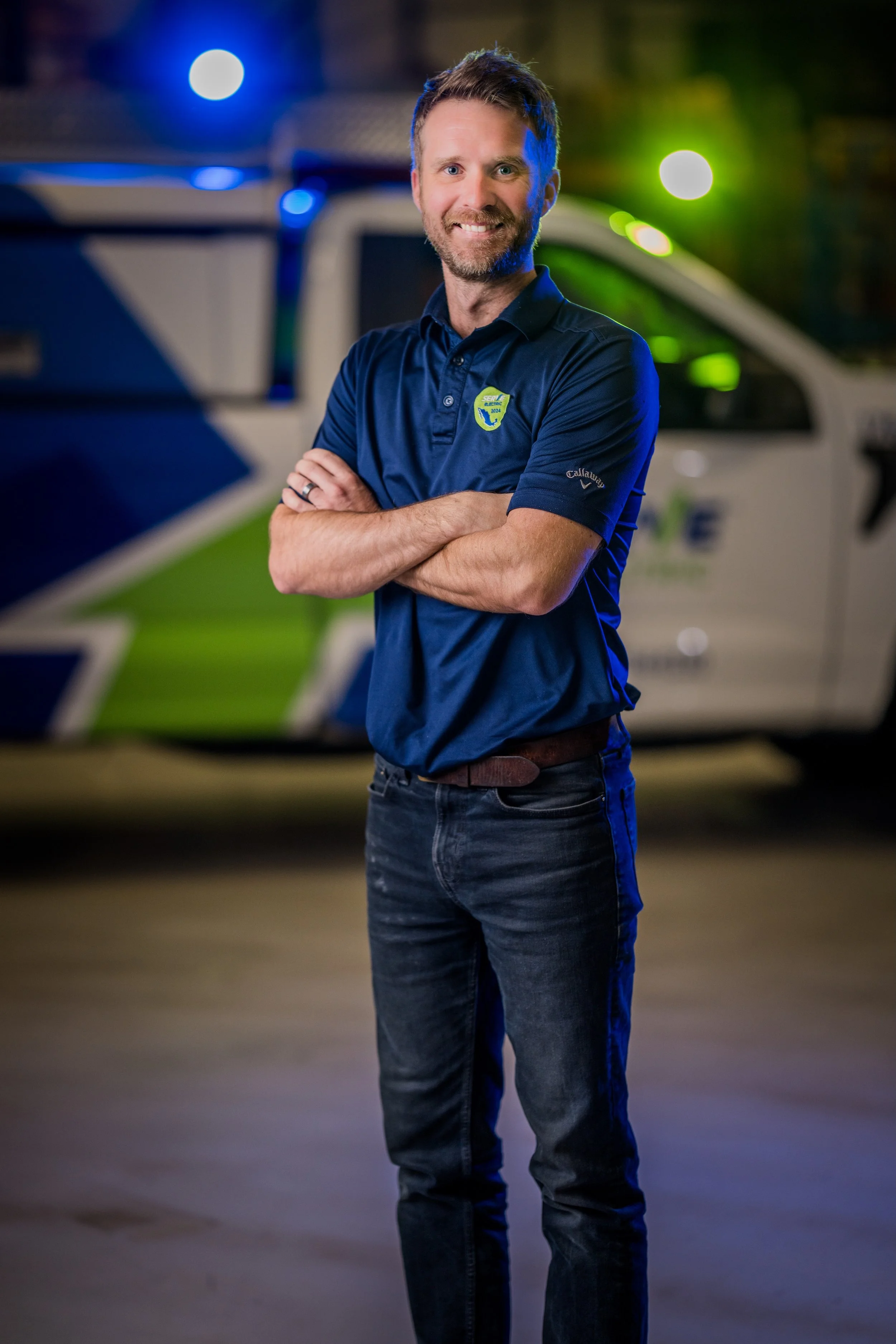 A man standing confidently with arms crossed in front of a police vehicle at night. He is wearing a navy blue polo shirt with a badge and the brand name 'Callaway' on the sleeve, smiling at the camera.