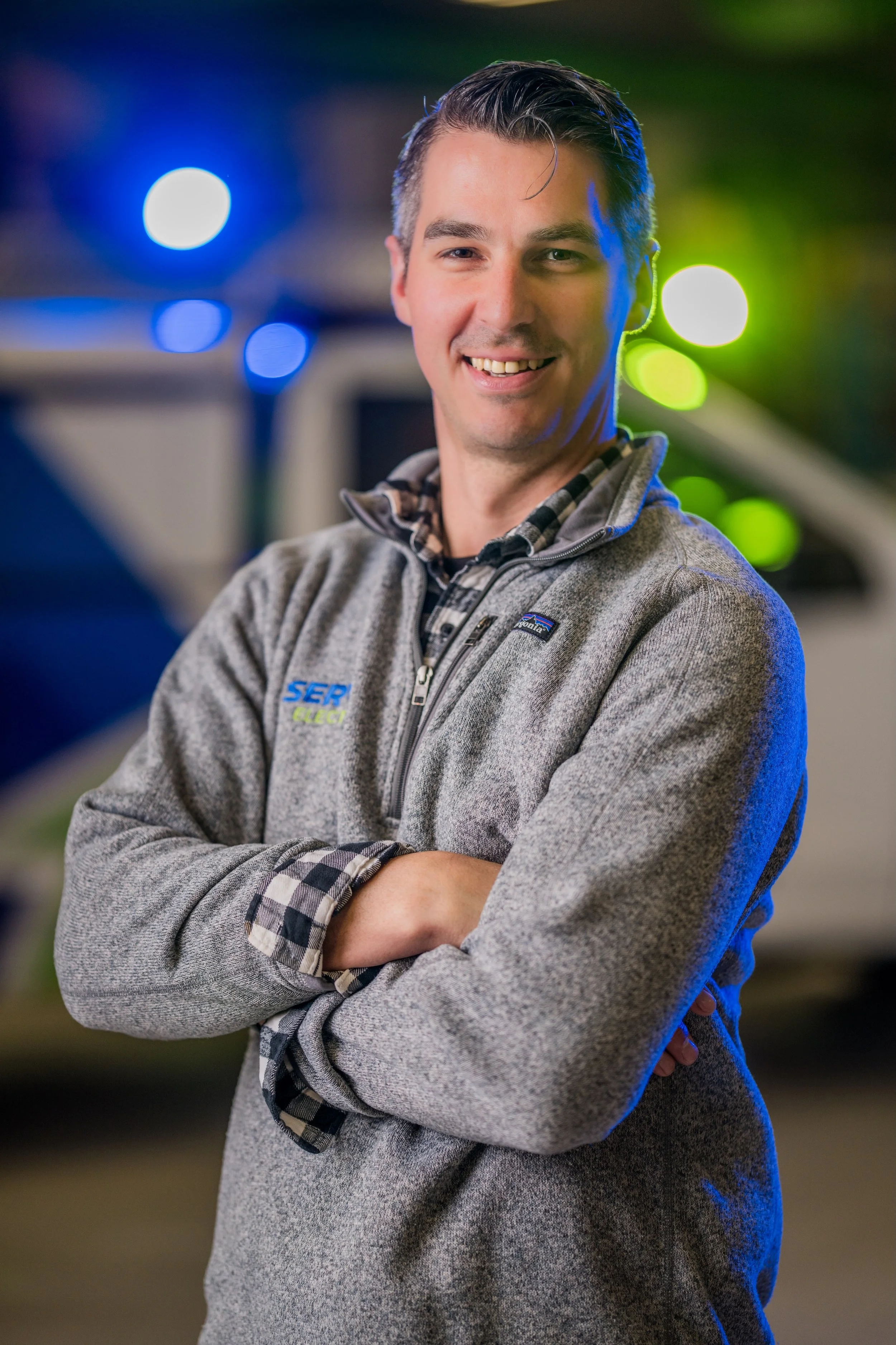 A man with short dark hair, wearing a gray Patagonia fleece jacket, standing outdoors at night with colorful lights in the background.