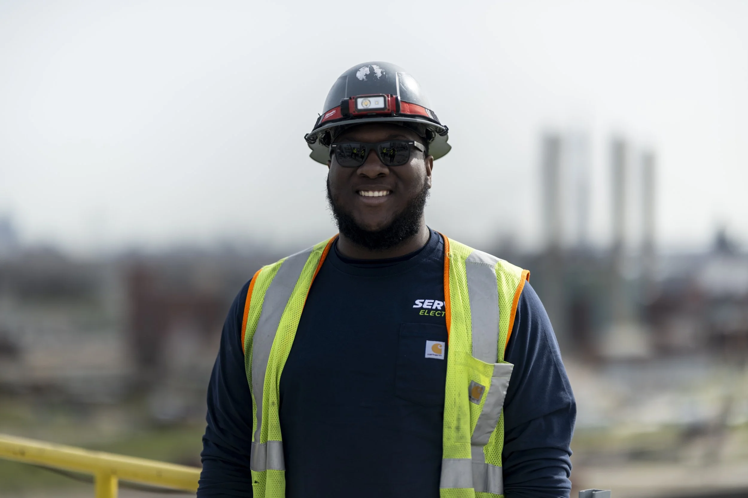 A smiling African American construction worker wearing a safety helmet, sunglasses, a dark blue shirt, and a yellow reflective safety vest on a construction site with a blurred cityscape in the background.