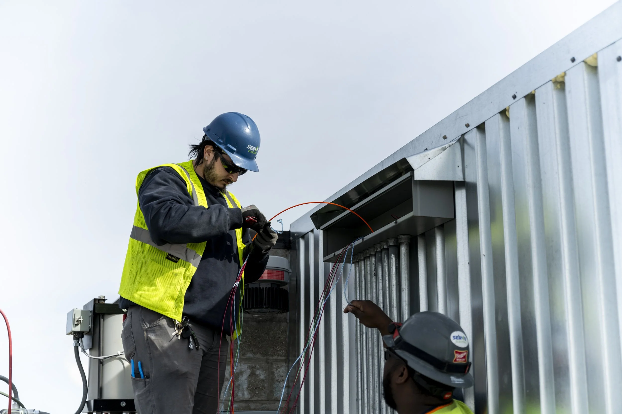 Two electrical workers wearing safety gear and helmets installing or repairing wiring on a metal building exterior.