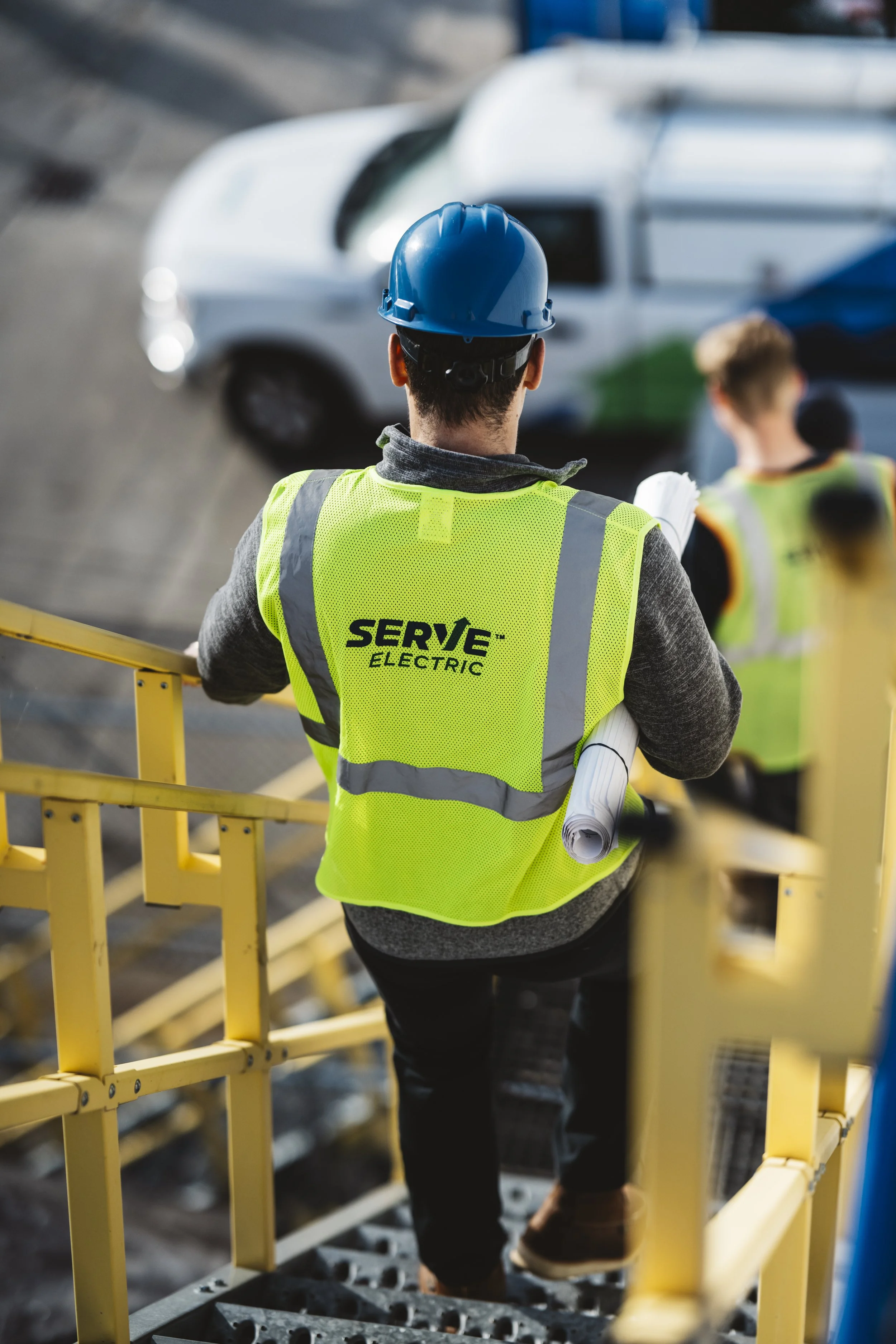 A worker wearing a blue hard hat and a bright yellow safety vest labeled 'SERVE ELECTRIC' is holding rolled-up blueprints while standing on a yellow staircase platform, overseeing a construction site with a white vehicle in the background.