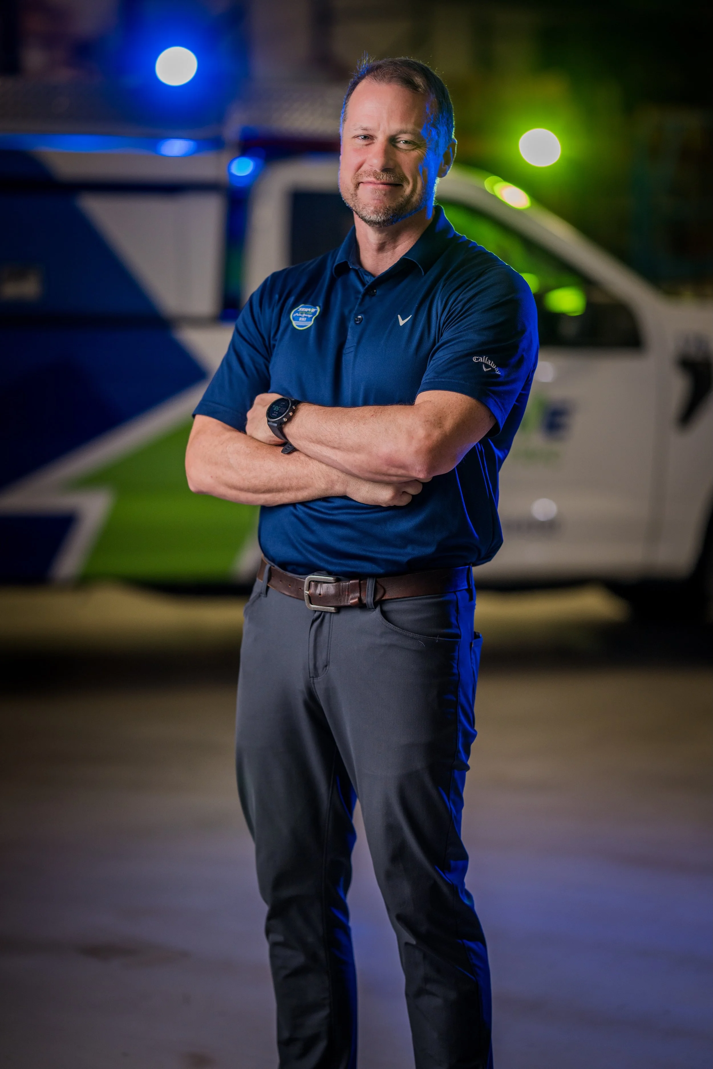A man in a blue polo shirt with a badge and logo stands with arms crossed in front of a police vehicle at night.