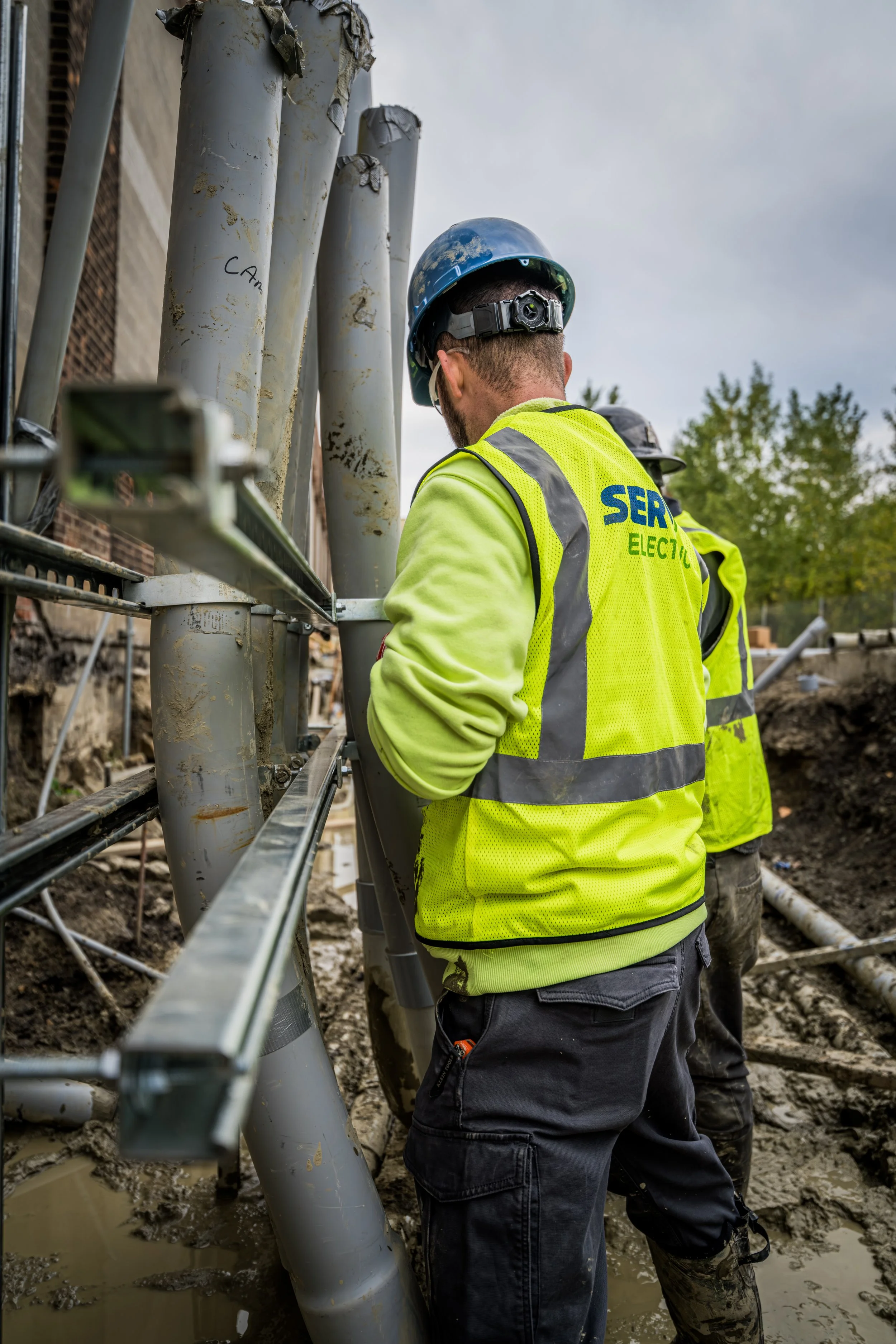 Construction workers in high-visibility jackets and helmets working on electrical or communication poles at a muddy construction site.