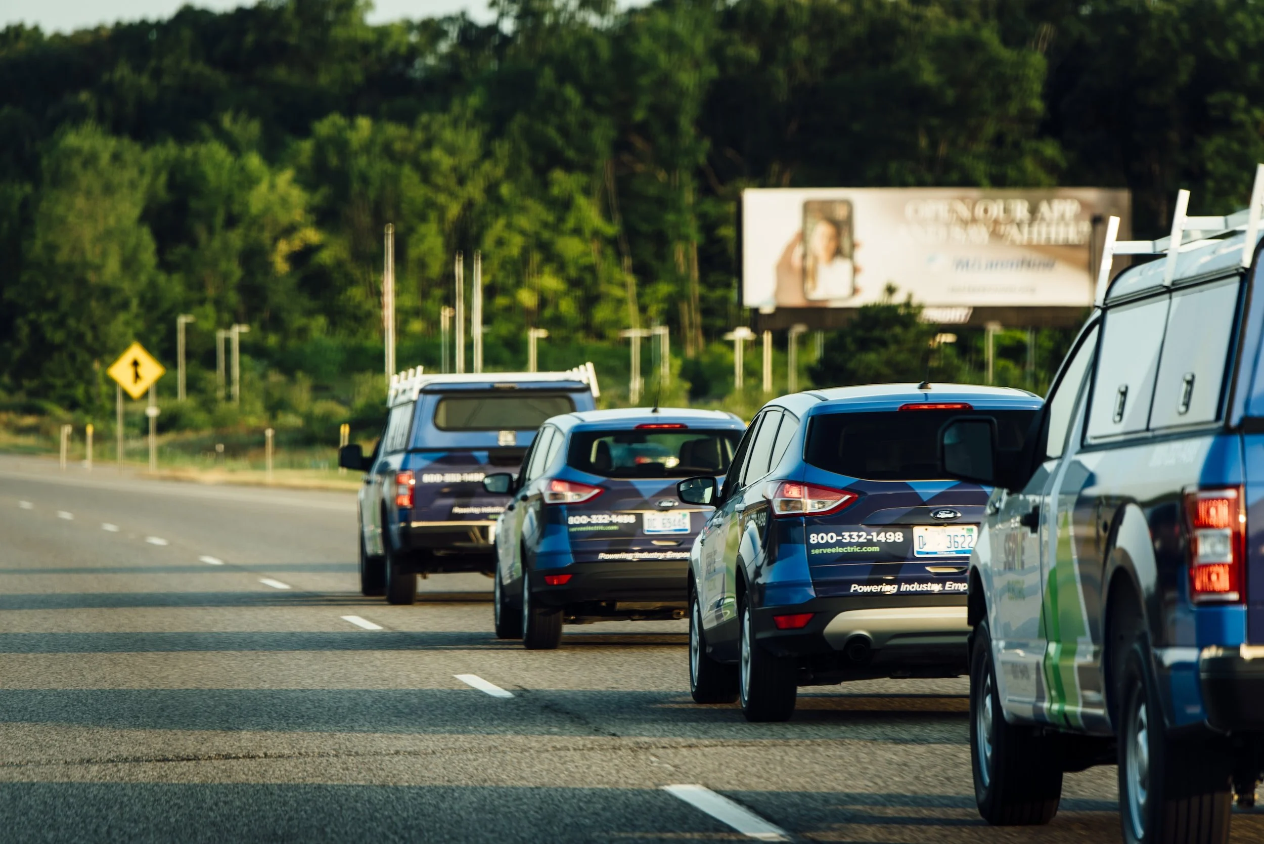 Line of four cars on the highway, with a forest in the background, and a billboard on the right side.