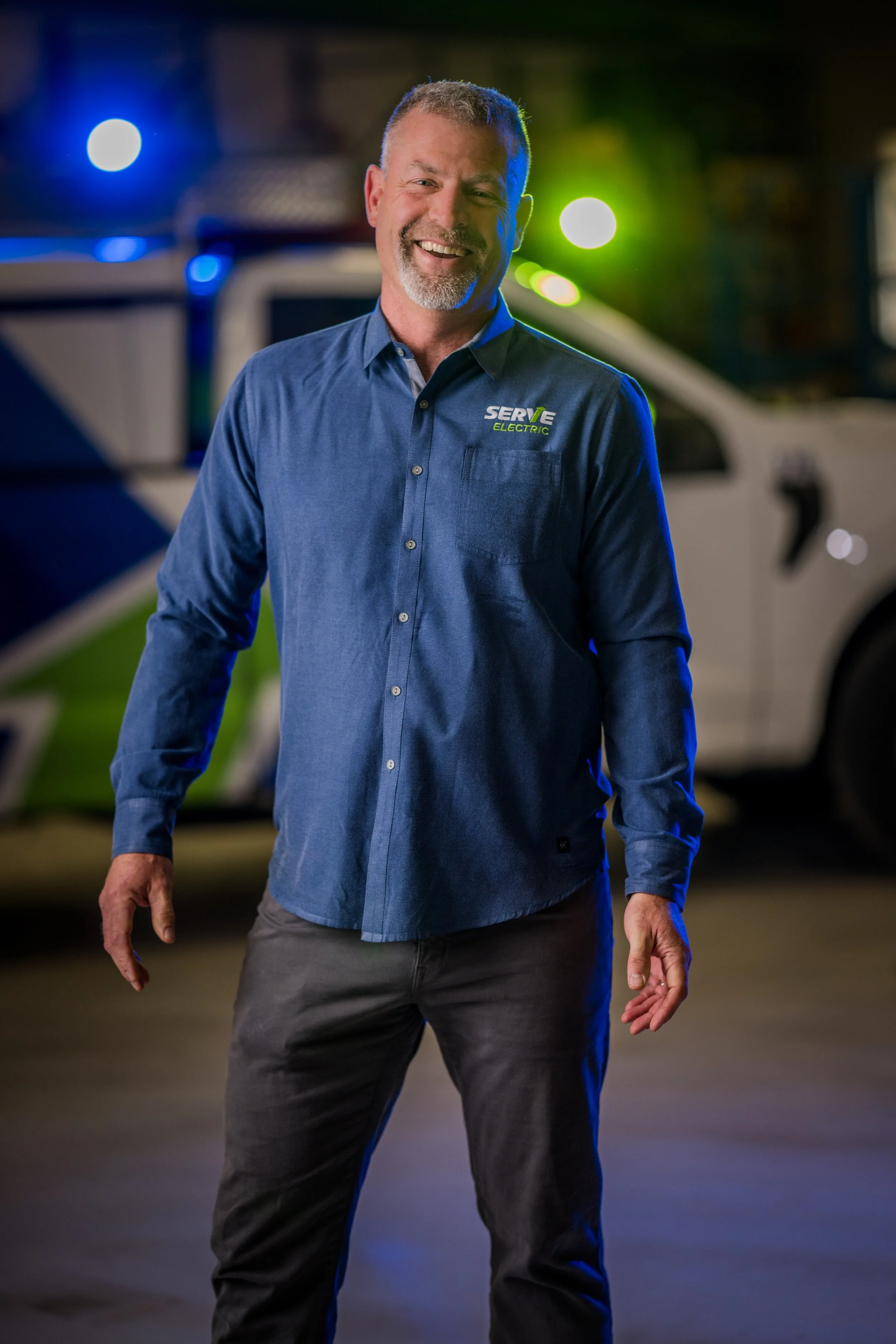 A smiling man in a blue shirt with the logo 'SERVE ELECTRIC' standing in front of a police vehicle at night.