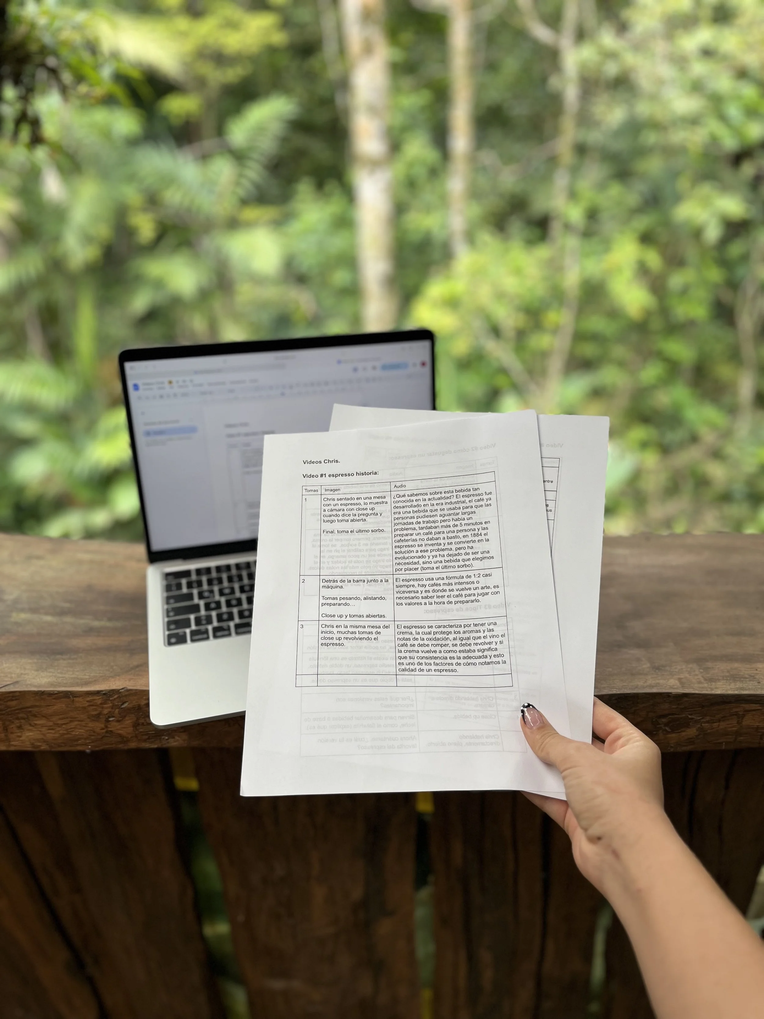 Una mano sostiene varias hojas impresas con texto, con un fondo de naturaleza con árboles verdes y un laptop abierto en una mesa de madera.
