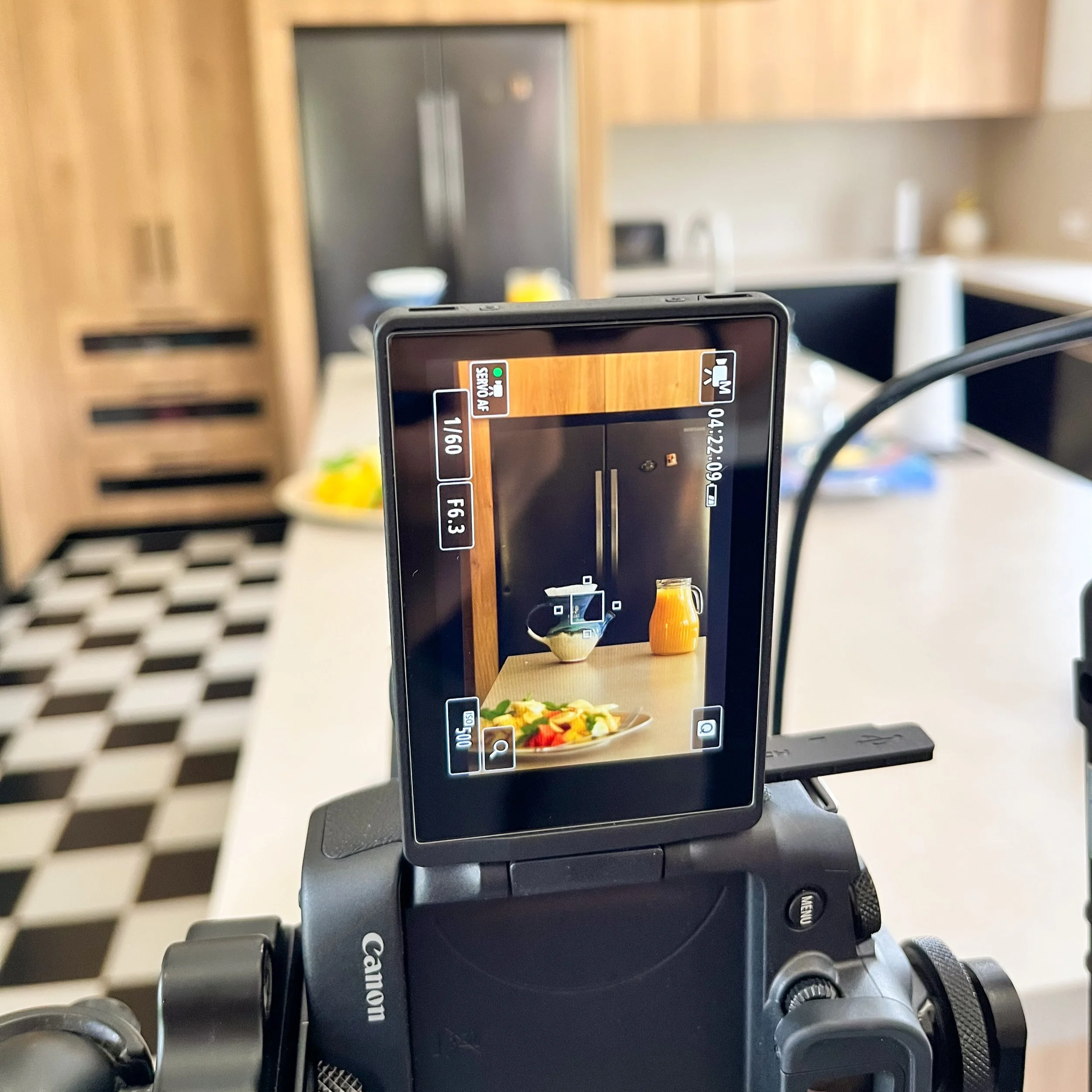 Vista de una cámara de video capturando una mesa con ensalada, una jarra con jugo y un recipiente con crema, en una cocina moderna con paredes de madera y suelo en patrón de cuadros blancos y negros.