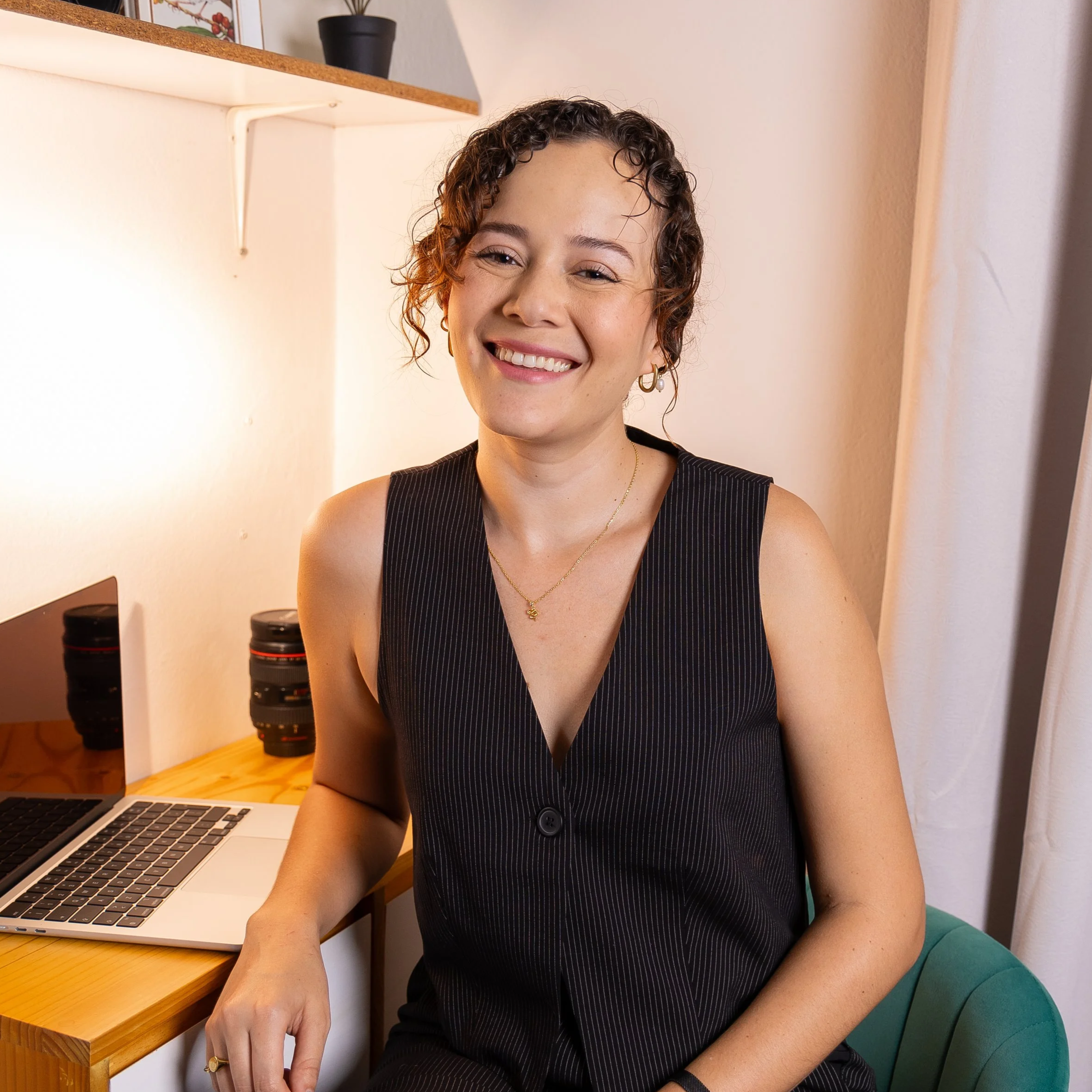 Mujer sonriendo, sentada en una silla, con una laptop y lentes en una mesa de madera a su lado, fondo con estantería, una planta y cortinas blancas.