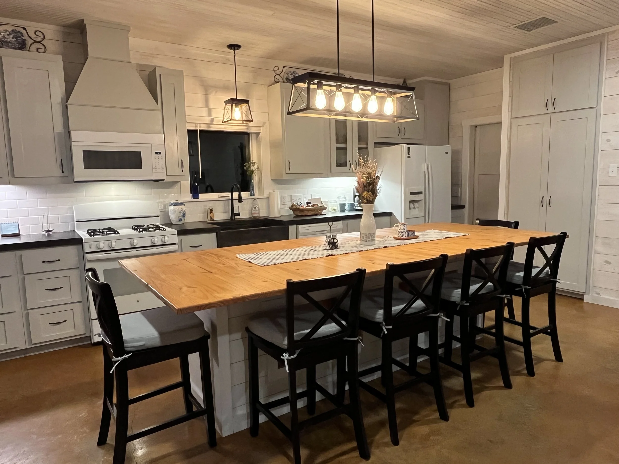 A spacious kitchen with a large wooden dining table surrounded by six black chairs. There are light fixtures hanging above the table, and the kitchen features white cabinetry, a white stove, microwave, refrigerator, and a black sink. The countertops 
