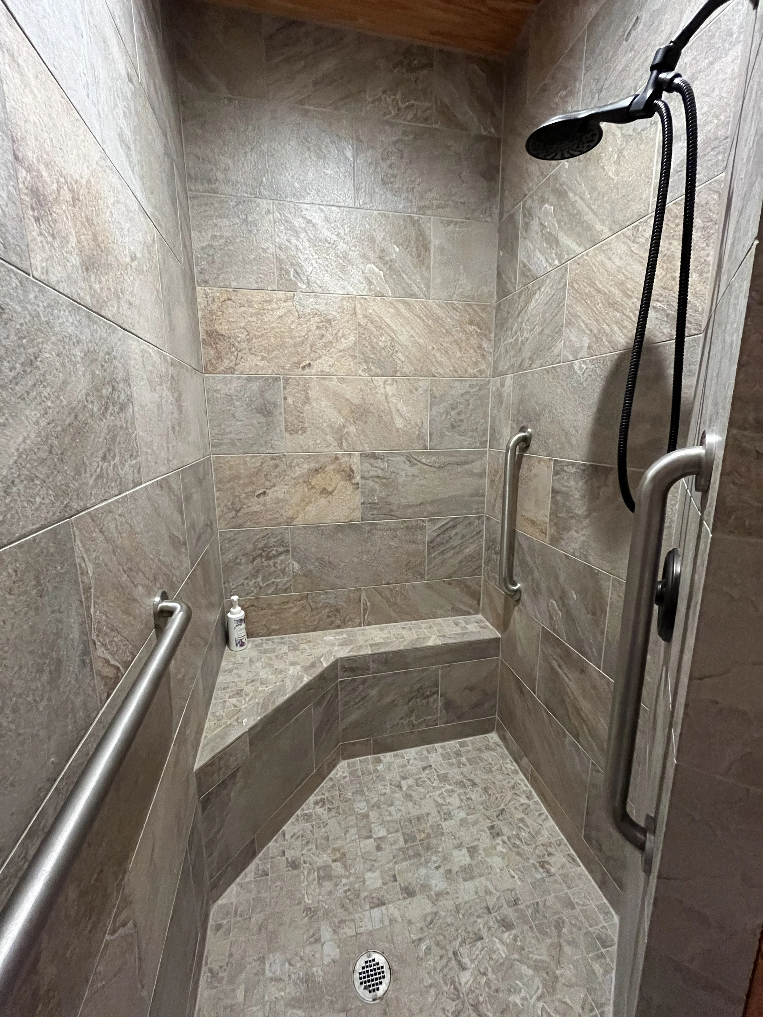 A walk-in shower with tiled walls and floor, a showerhead, two grab bars, a built-in seat in the corner, and a bottle of soap on the seat.