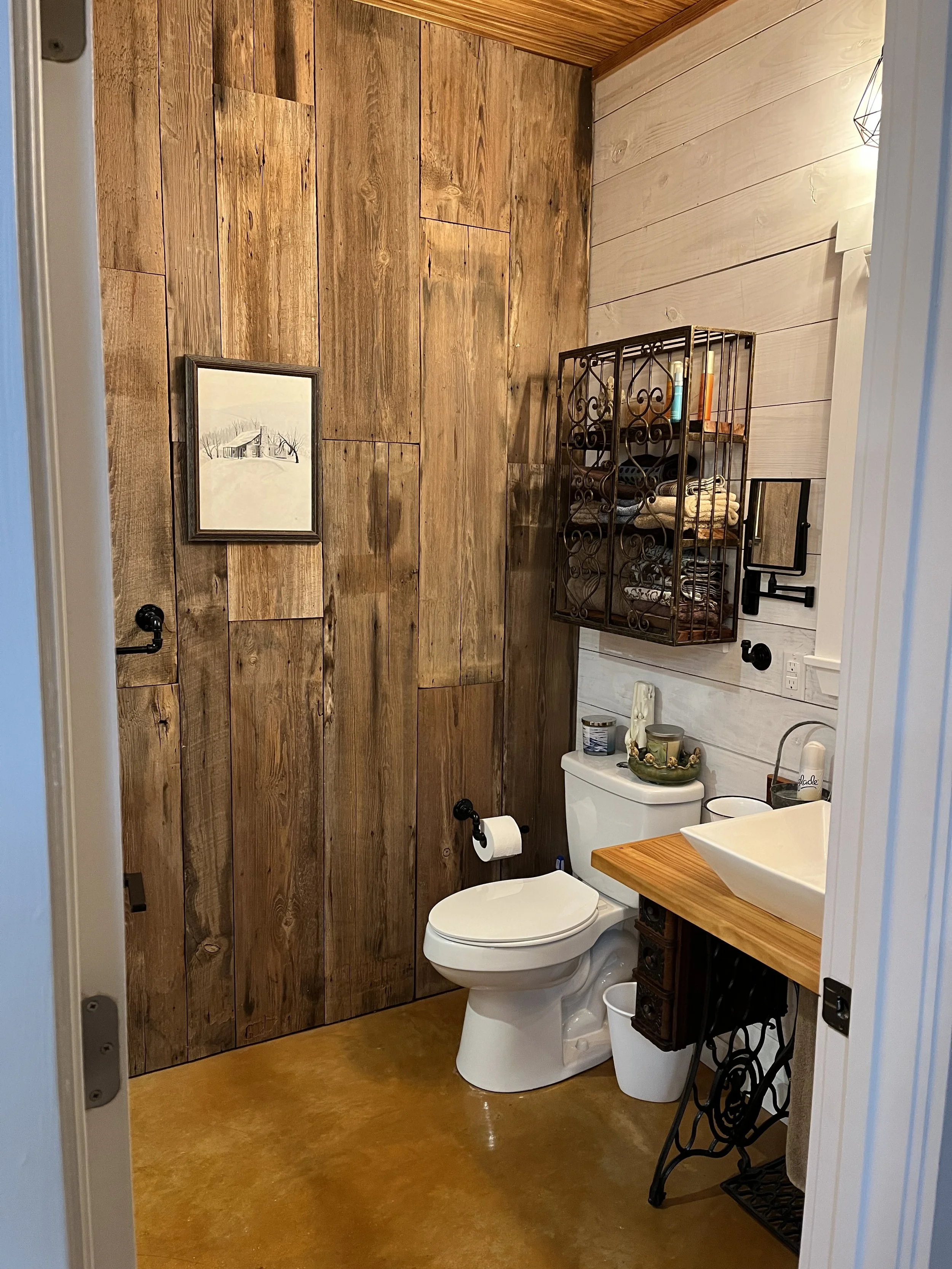 A small bathroom with a wooden wall, a white toilet, a wooden vanity with a white vessel sink, a wall-mounted iron shelf with folded towels and toiletries, and a framed picture on the wall.