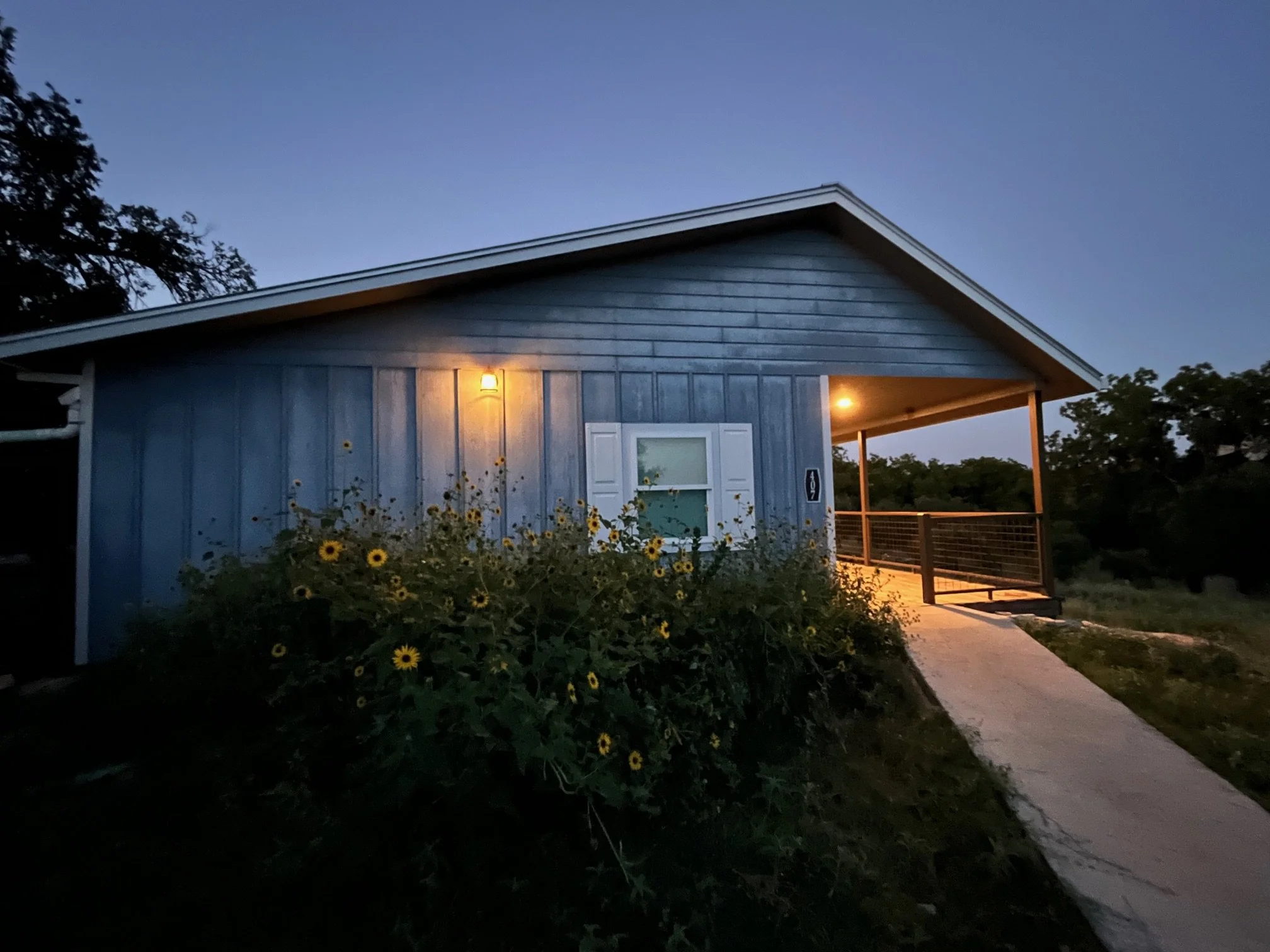 A blue house with white shutters, a front window, and a porch surrounded by flowers at dusk. The house has exterior lighting and a pathway leading to the entrance.