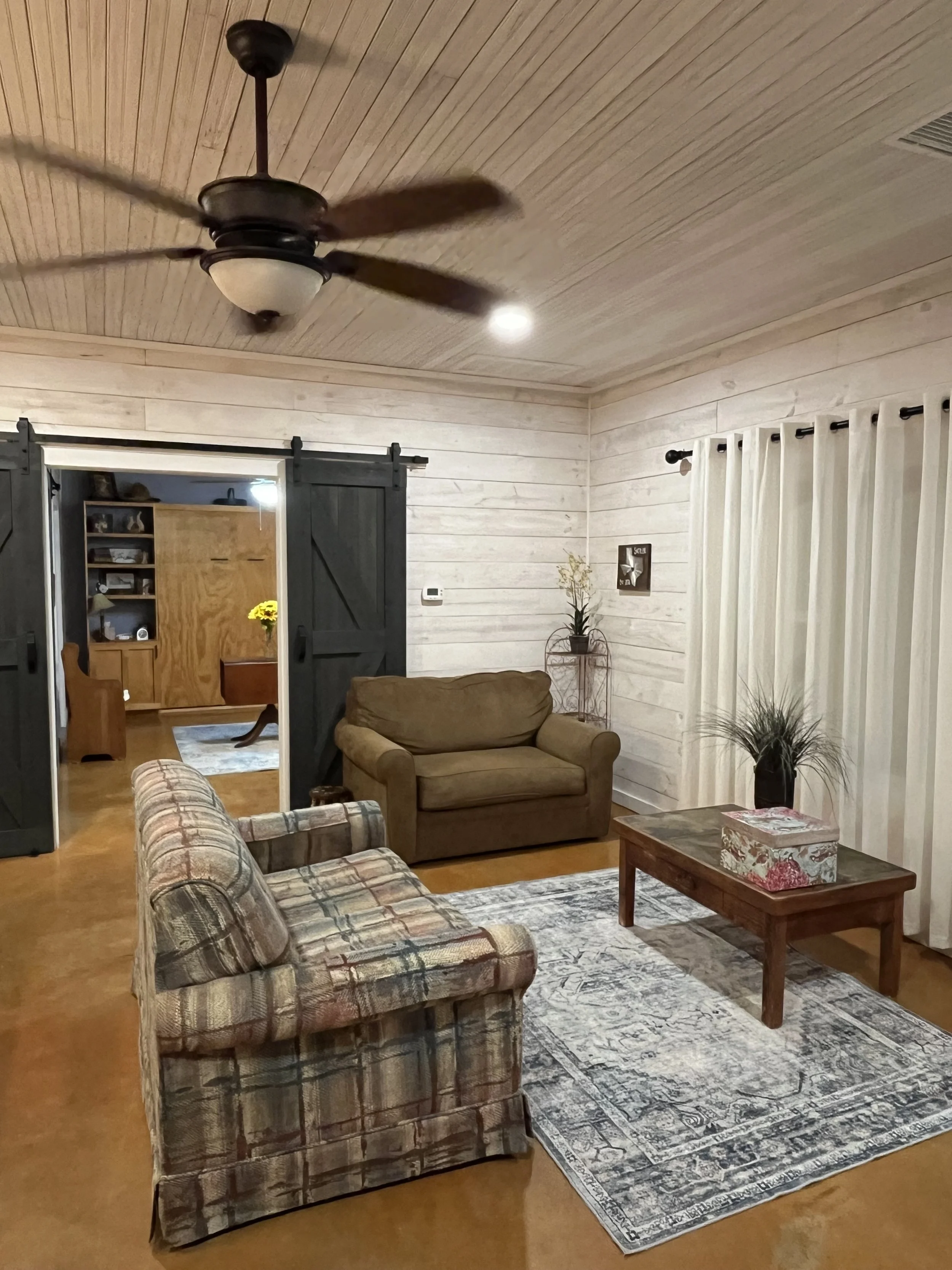 Living room with two sofas, a wooden coffee table, a decorative rug, and curtains. In the background, there is a doorway with black sliding barn doors revealing another room with shelves and flowers.