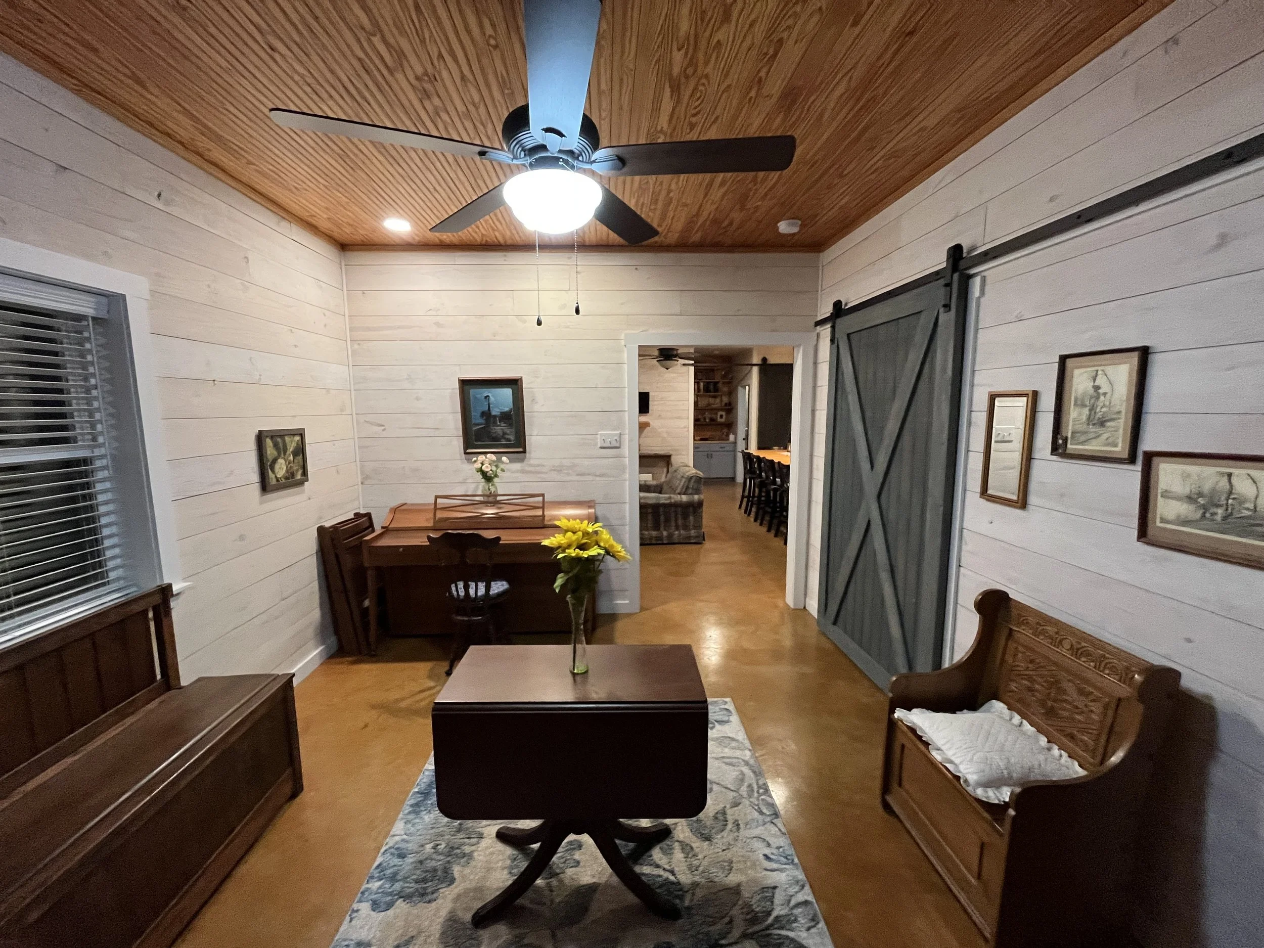 Living room with white wooden walls and a wood-paneled ceiling, ceiling fan and light fixture, window with blinds, various paintings on the walls, wooden furniture including a bench, a small table with a sunflower, a dining table with chairs, and an 