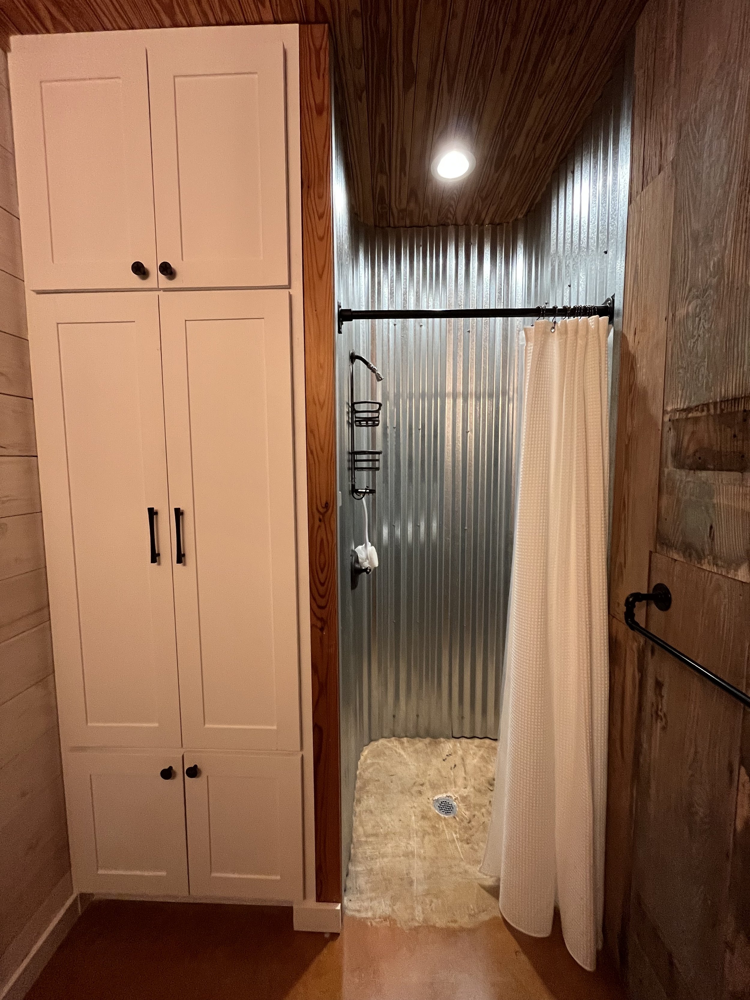 A walk-in shower with corrugated metal walls, a black curtain rod with a beige curtain, a showerhead, and built-in shelving. Adjacent to the shower is white cabinetry and wood-paneled walls, with a small rug on the floor.