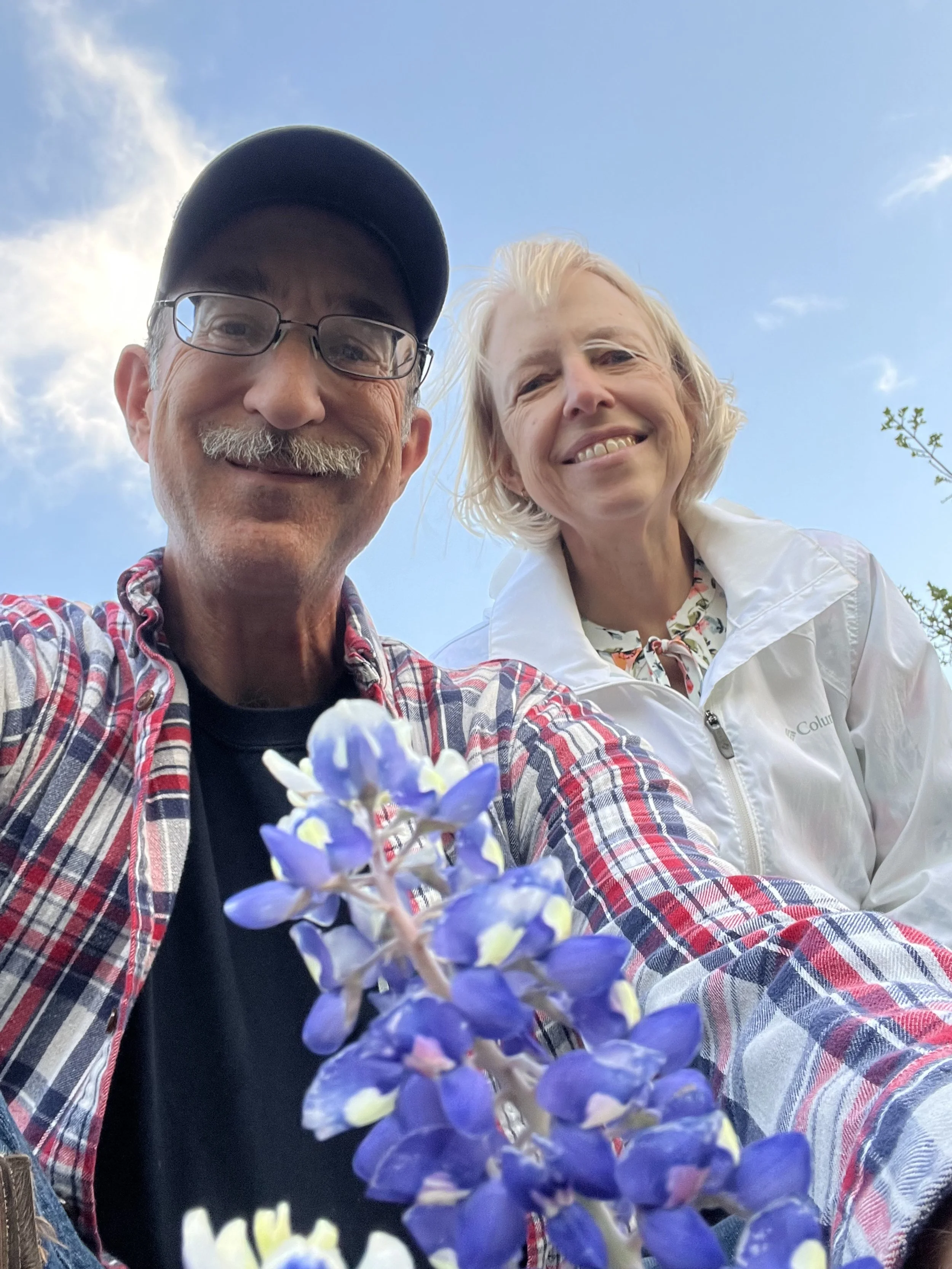 A man and a woman standing outdoors during daytime, smiling at the camera. The man wears glasses and a baseball cap, and holds a cluster of purple and white flowers. The woman has short blonde hair and wears a light-colored jacket.