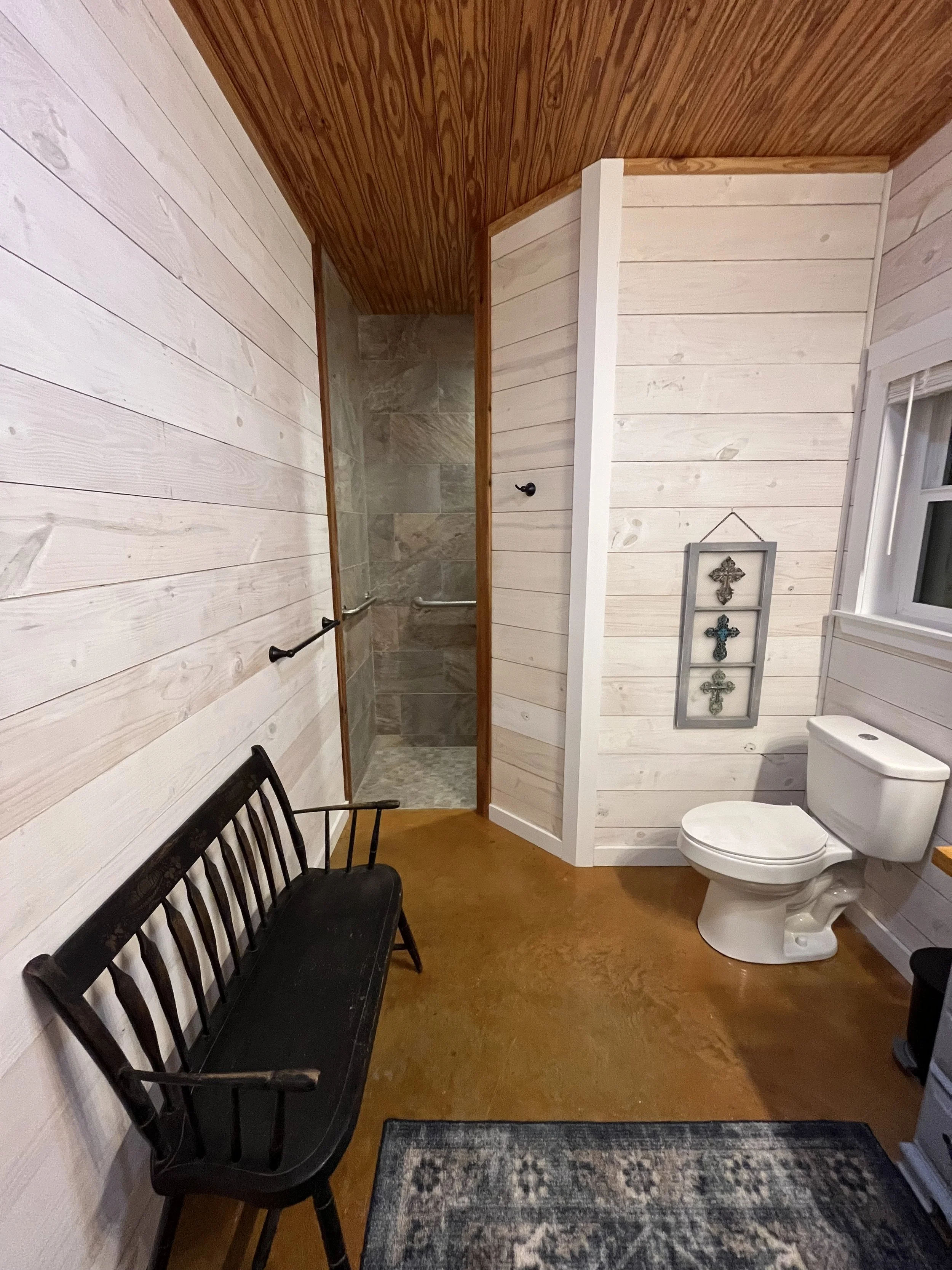 A rustic bathroom with white-paneled walls, a wooden ceiling, a black bench, a patterned rug, a toilet, a window, and a shower area with stone tiles and grab bars.