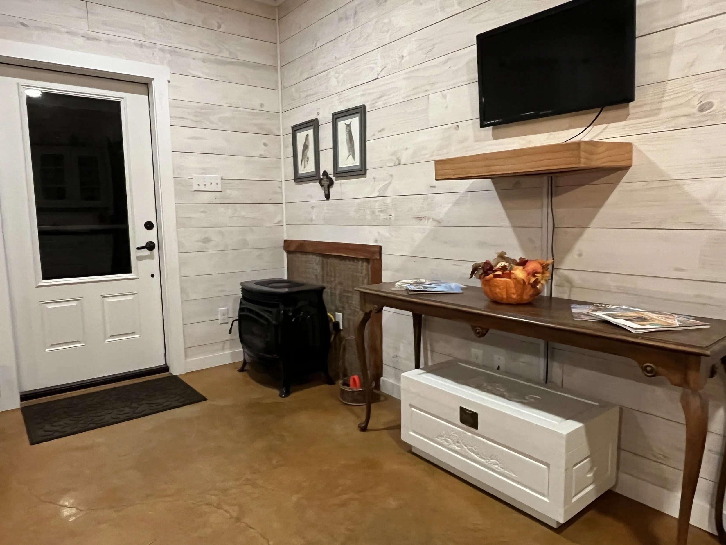 Entryway with a white door, black doormat, wooden wall, framed bird pictures, mounted TV, wooden shelf, beige table with a plant basket, magazines, and a white storage bench underneath.