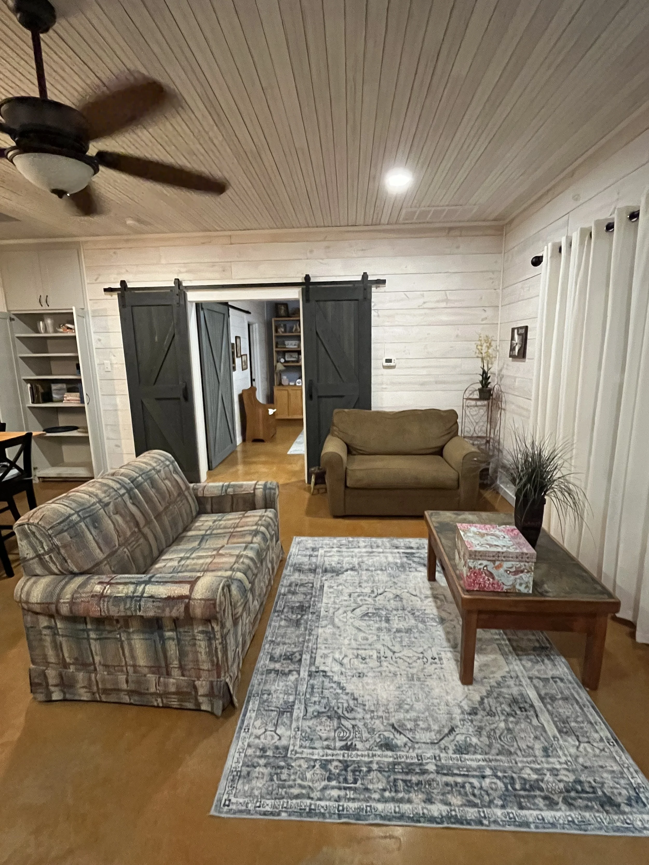 Cozy living room with plaid sofa, green armchair, wooden coffee table, patterned rug, plants, and white-paneled walls with sliding barn doors.