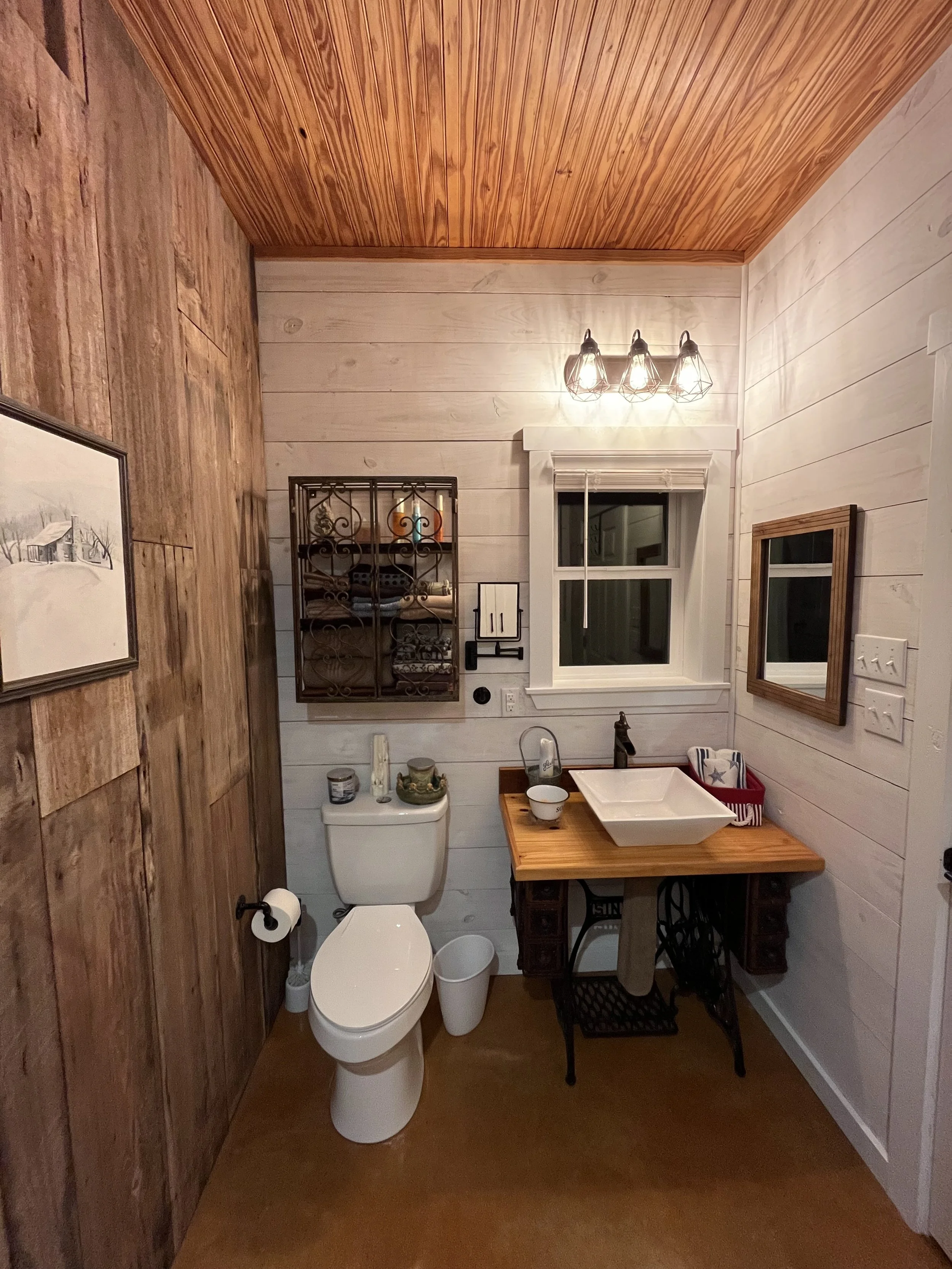 A rustic bathroom with mixed wood paneling, a small sink with a wooden countertop, a toilet, and various decor items.