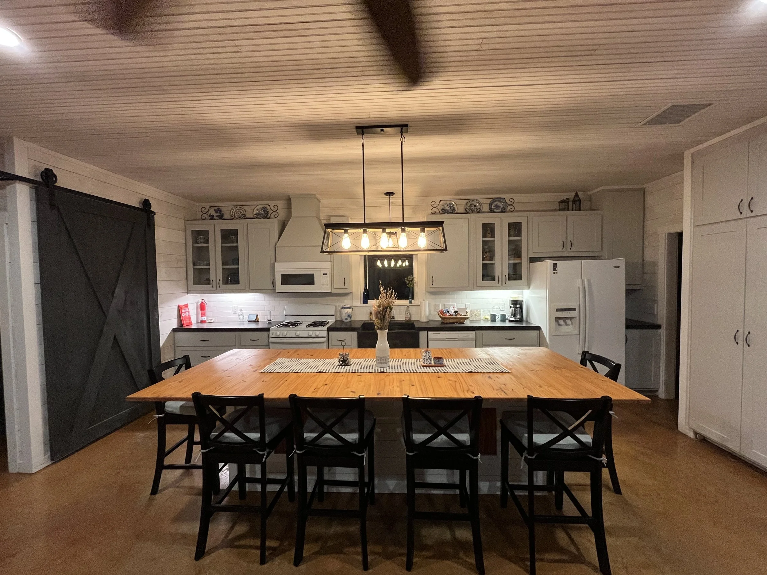 A farmhouse-style kitchen with a large wooden dining table with six black chairs, white cabinetry, a white stove with a microwave above, a white refrigerator, and a black countertop. An industrial-style pendant light hangs above the table.