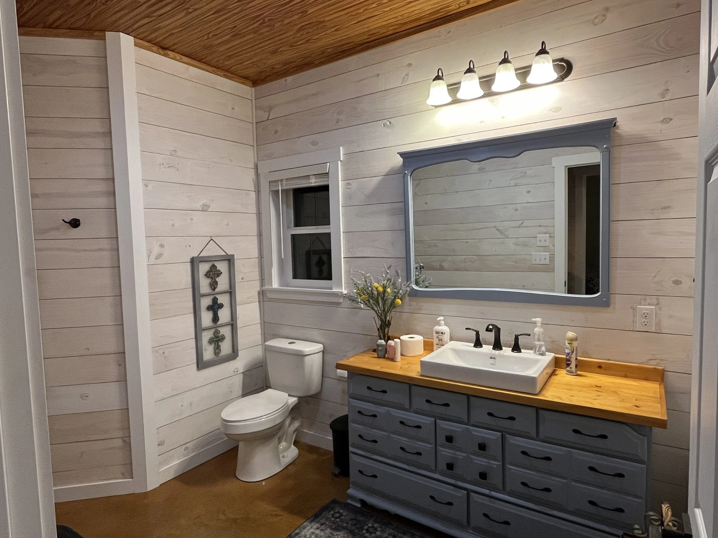 Bathroom with white wood-paneled walls, a white toilet, a blue dresser with a wooden countertop, a rectangular sink with black fixtures, a large mirror, a window, and four light fixtures above the mirror.
