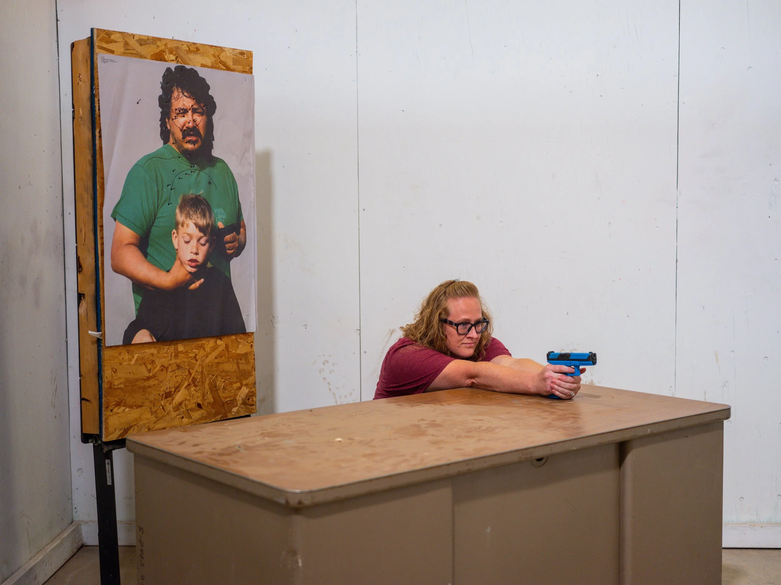 A woman with glasses, wearing a maroon shirt, is seated at a wooden table aiming a blue handgun. To her side, there is a portrait of a man with curly hair and a beard, wearing a green shirt, holding a microphone, with a boy in front of him.