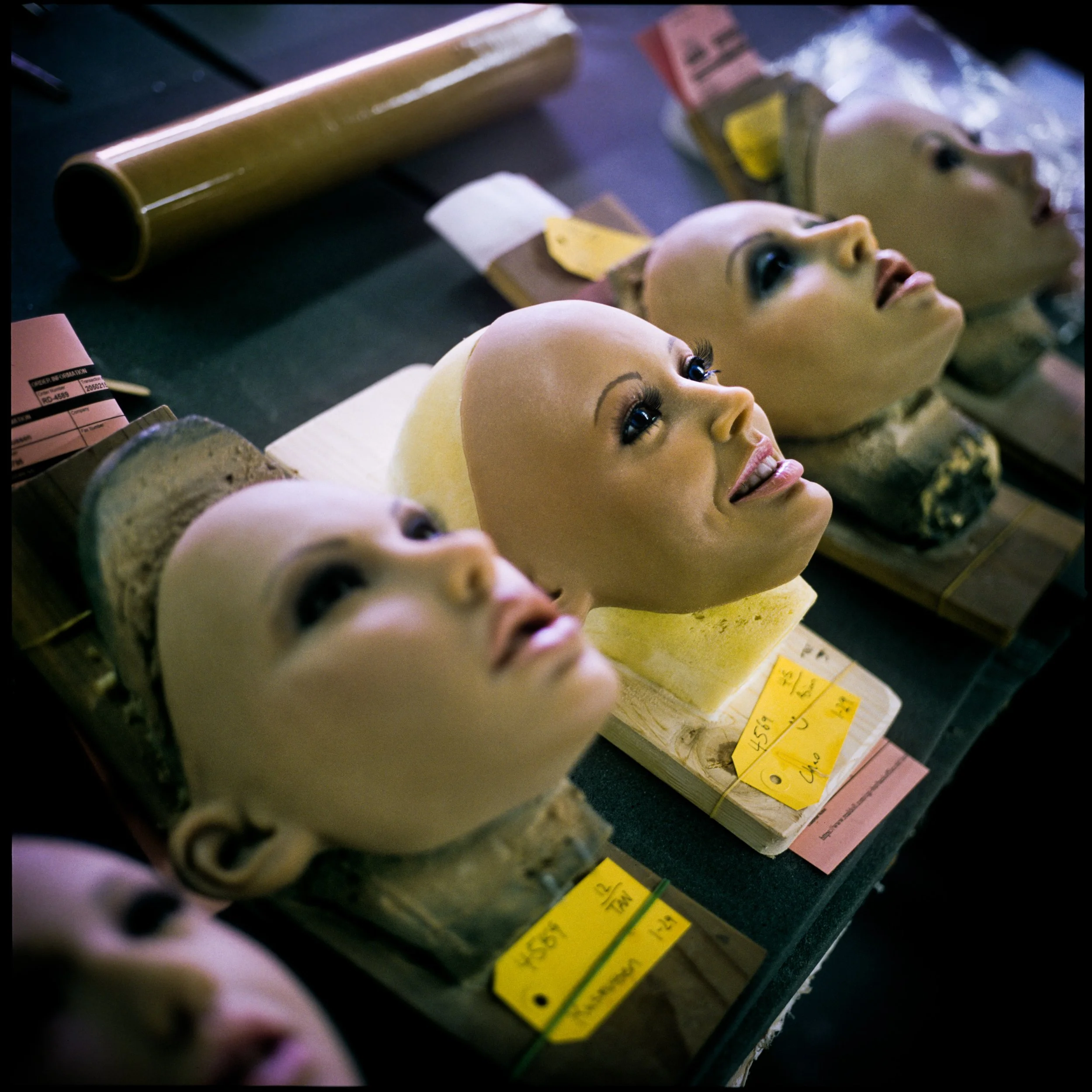 Four REAL DOLL heads with realistic faces, eyes, and makeup, placed on blocks on a workbench, with various crafting supplies around.