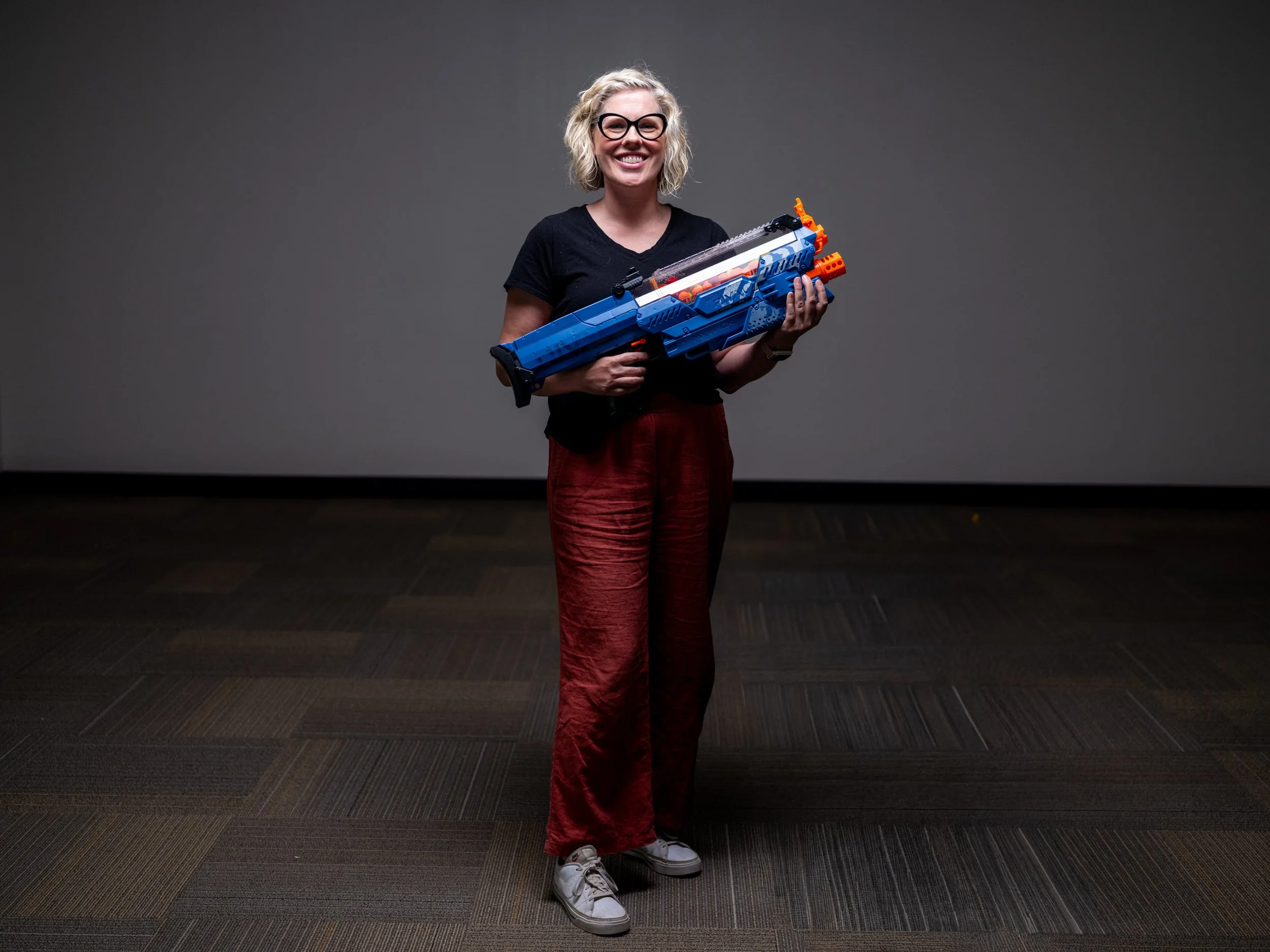 A woman with blonde curly hair, glasses, and a black shirt holding a large toy gun, smiling, standing in an empty room with a plain gray wall and patterned carpet from the HBO Documentary "Thoughts & Prayers"