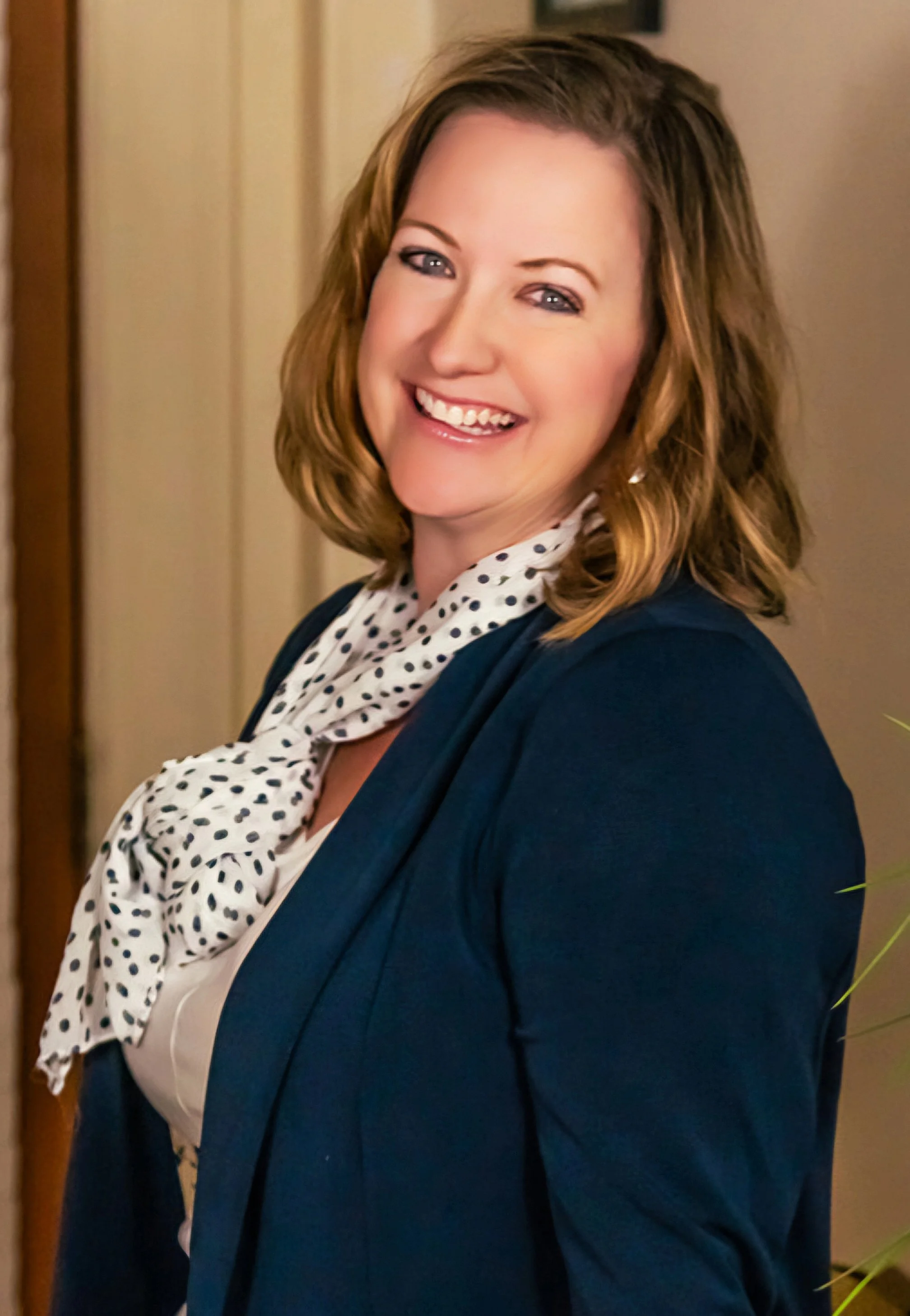 A woman with shoulder-length wavy light brown hair smiling, wearing a navy blazer, white blouse, and a white scarf with black polka dots, standing indoors with a beige wall and a wooden door in the background.