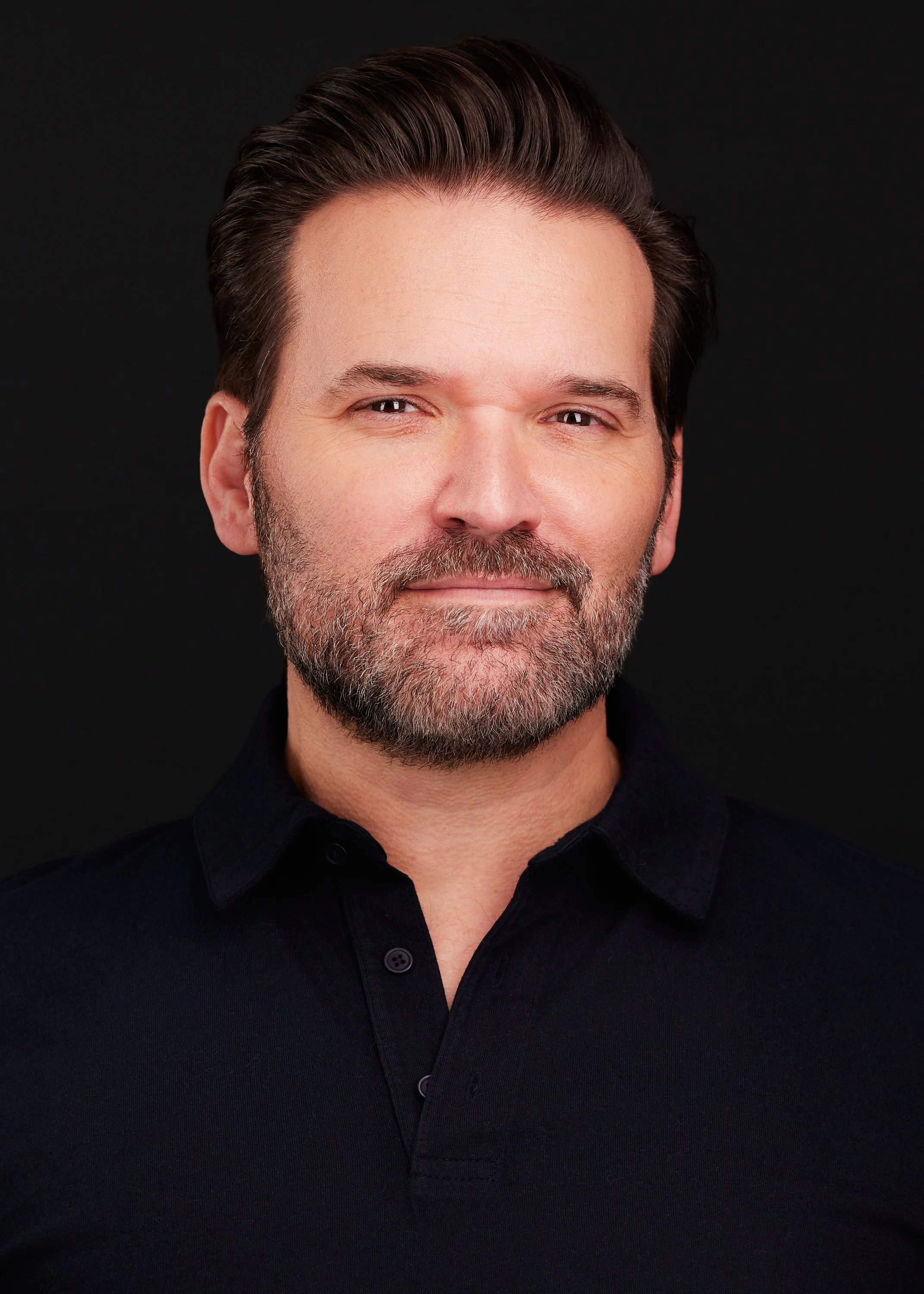 Close-up portrait of a middle-aged man with dark hair and a beard, wearing a black collared shirt, against a black background.