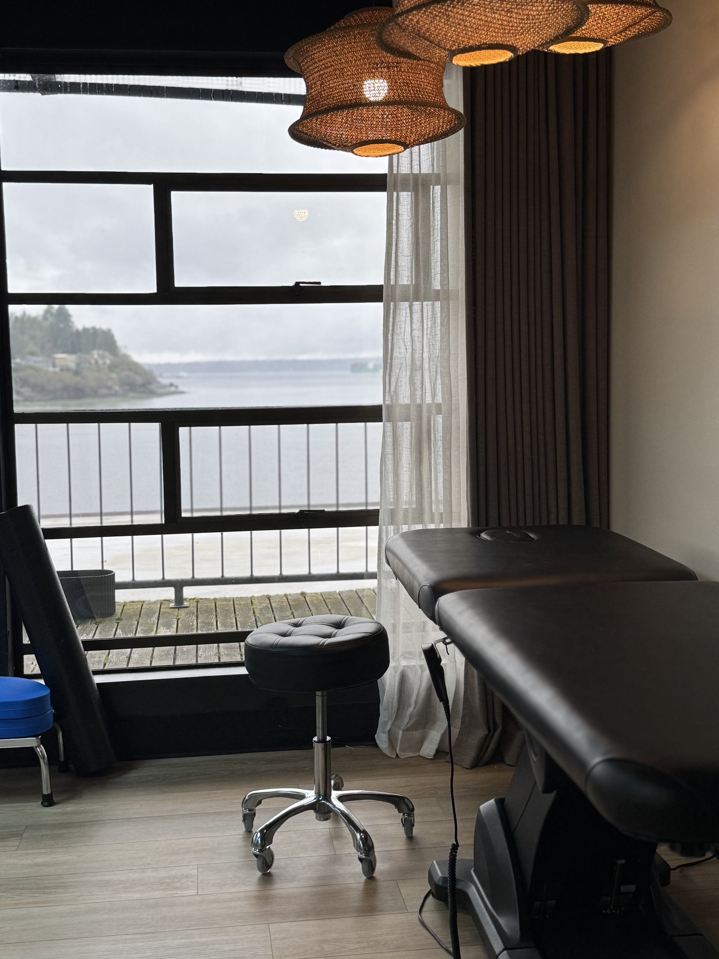 Interior of a massage room with a massage table, stool, and view of water outside a large window.