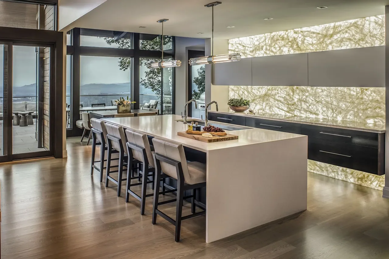 Modern kitchen with a white island, four beige chairs, two pendant lights, large windows with a view of trees and mountains, black lower cabinets, beige upper cabinets, and a decorative bowl of greenery.