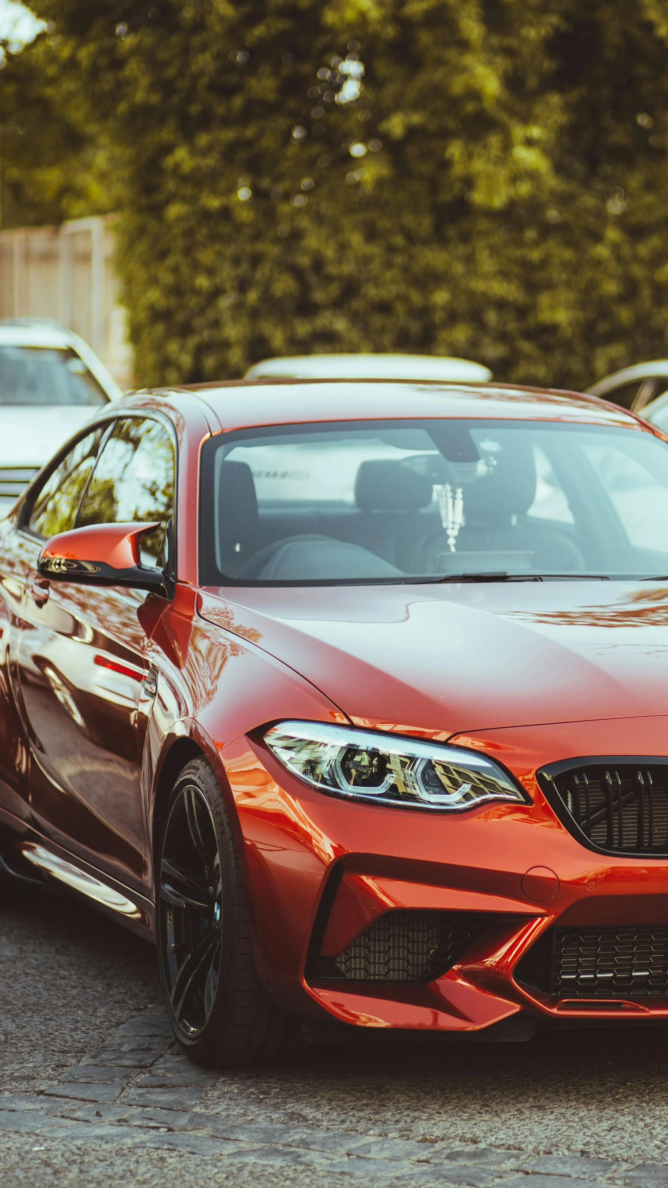 A close-up view of a red sports car parked on a street with a blurred background of trees and other vehicles.