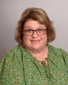 A woman with short, curly, light brown hair, wearing glasses and a green floral blouse, smiling at the camera.