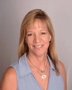 A woman with blonde hair wearing a light blue sleeveless top and jewelry, smiling at the camera.