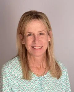 portrait of a woman with blonde hair, smiling, wearing a light blue and white patterned blouse, in front of a plain background.