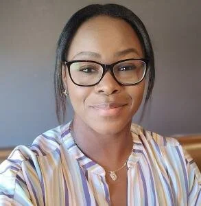 A woman with glasses and earrings, wearing a striped shirt, smiling at the camera in front of a gray wall.
