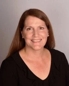 A woman with long brown hair smiling, wearing a black top, against a light gray background.