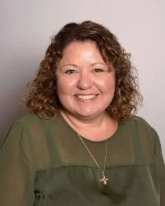 A woman with curly brown hair, smiling, wearing a green top and a cross necklace.