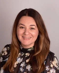 A woman with long, wavy brown hair smiling at the camera, wearing a black floral blouse against a plain white background.